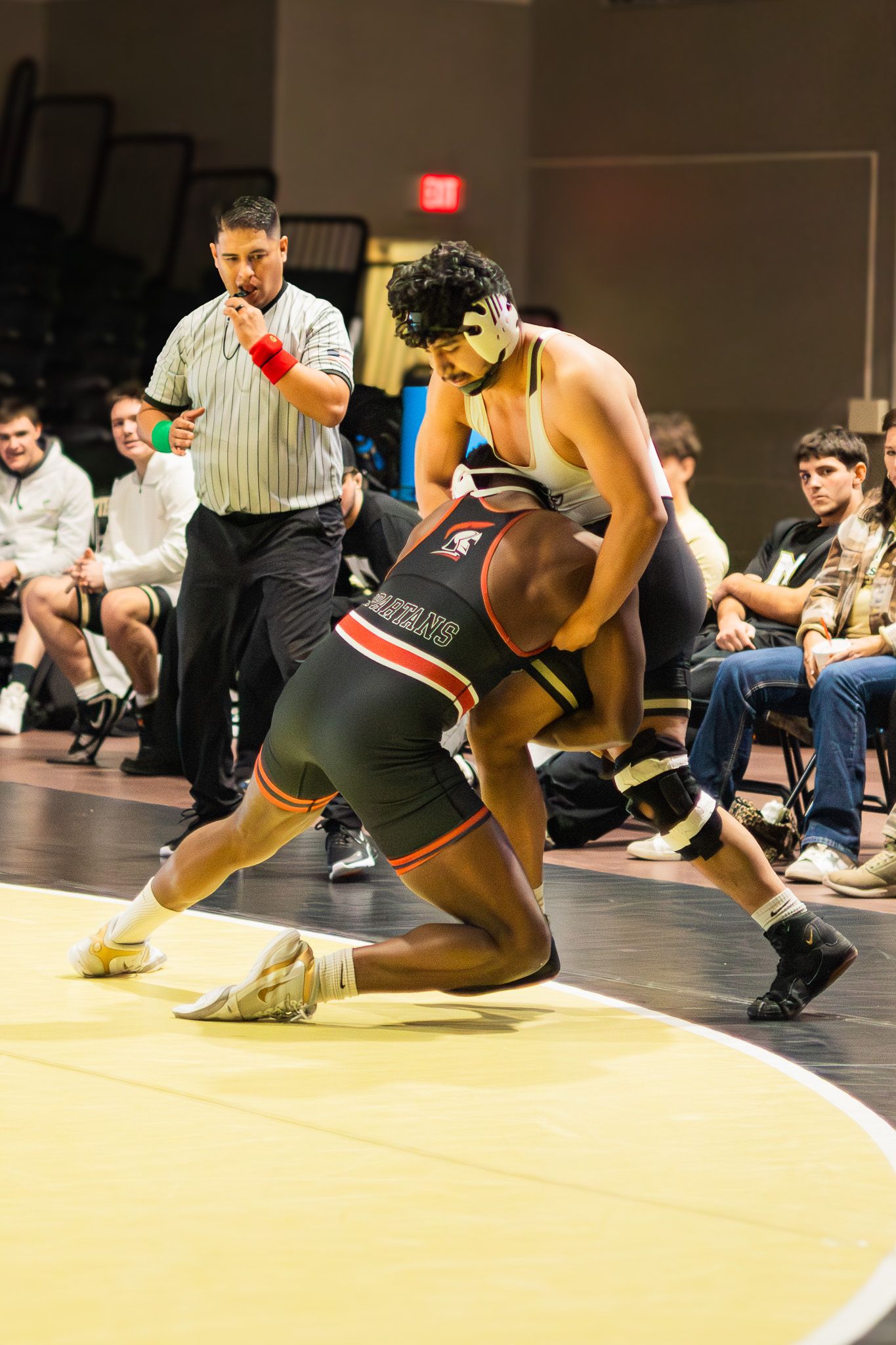 Referee watches closely as two wrestlers grapple during competitive college wrestling match.