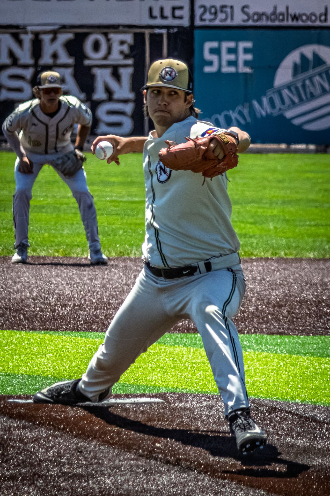 A baseball pitcher winds up to throw during a game with a fielder positioned in the background.