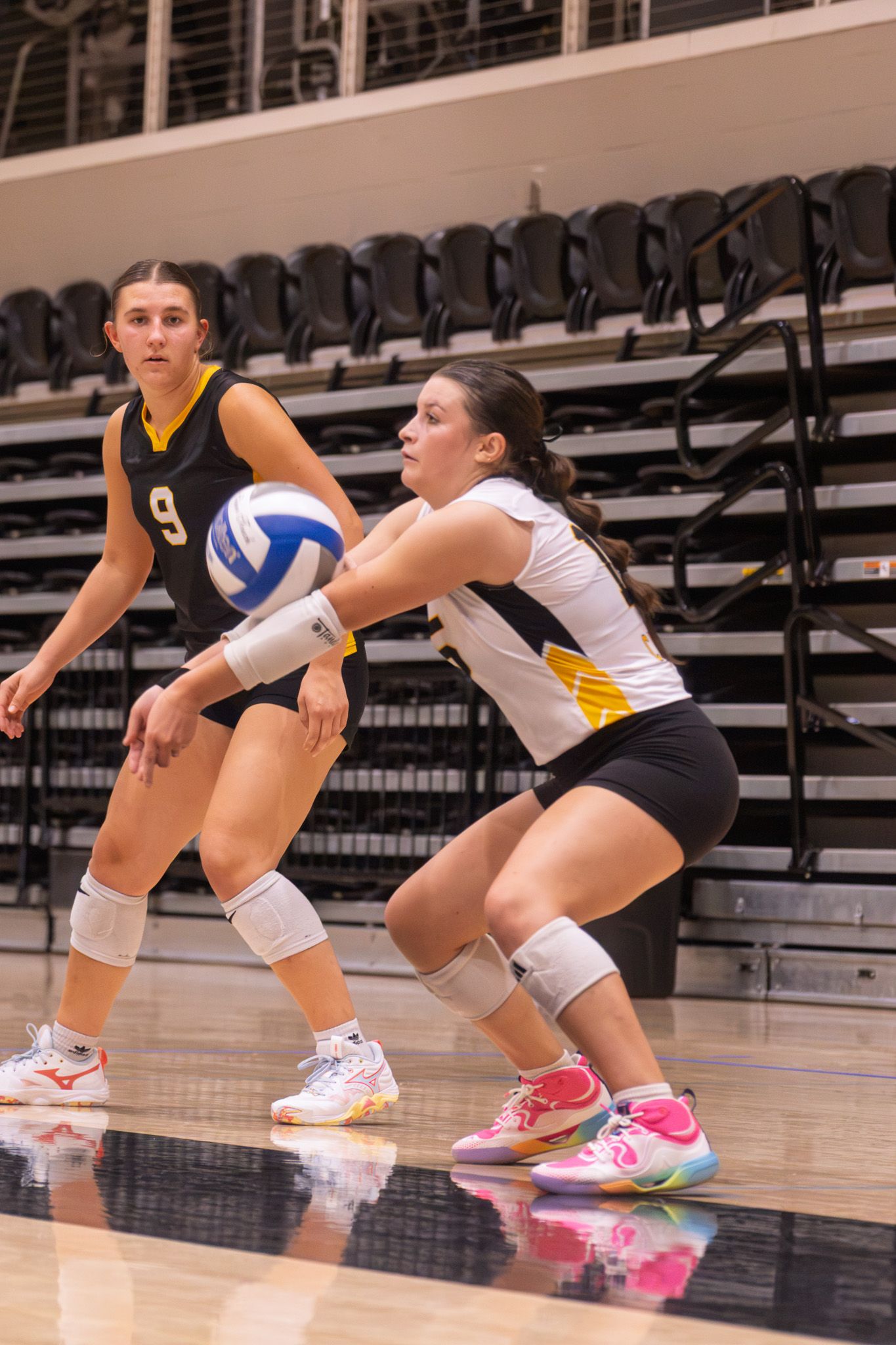 Female volleyball player performs a forearm pass dig while teammate number 9 watches during indoor match.