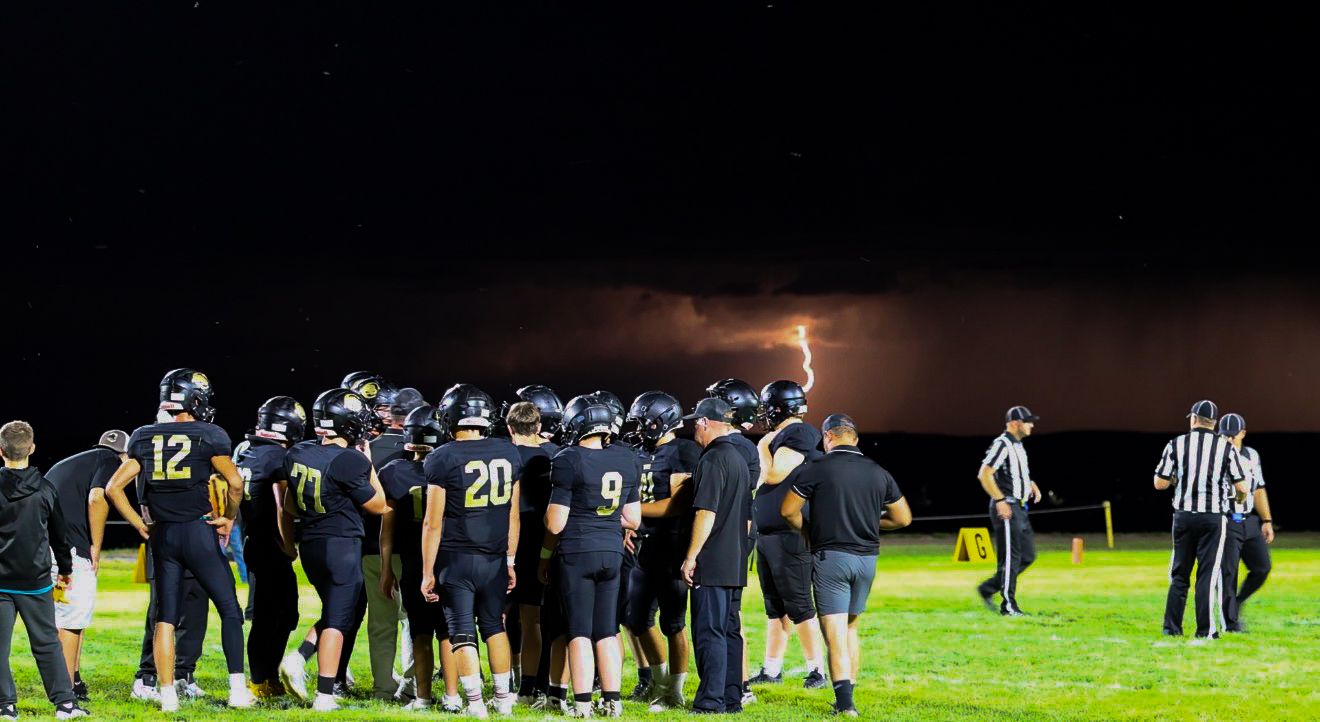 High school football team huddles on field during a dramatic night game with lightning in background.