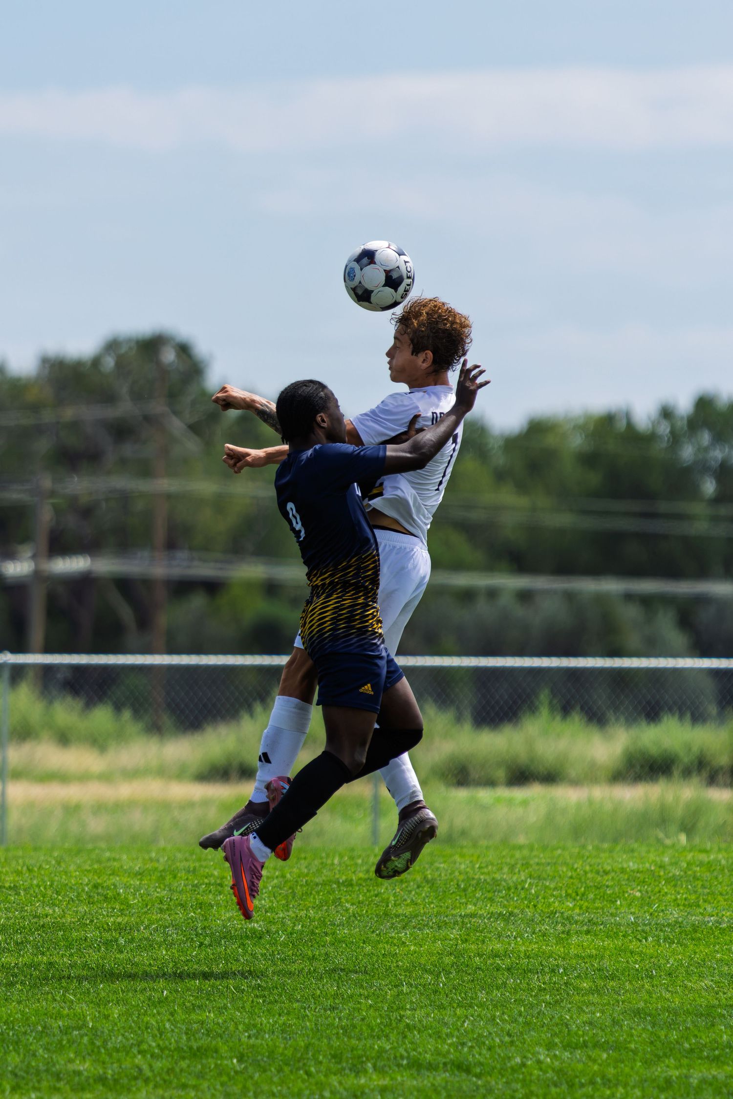 Two soccer players jump to head the ball during an intense aerial duel on a green grass field.