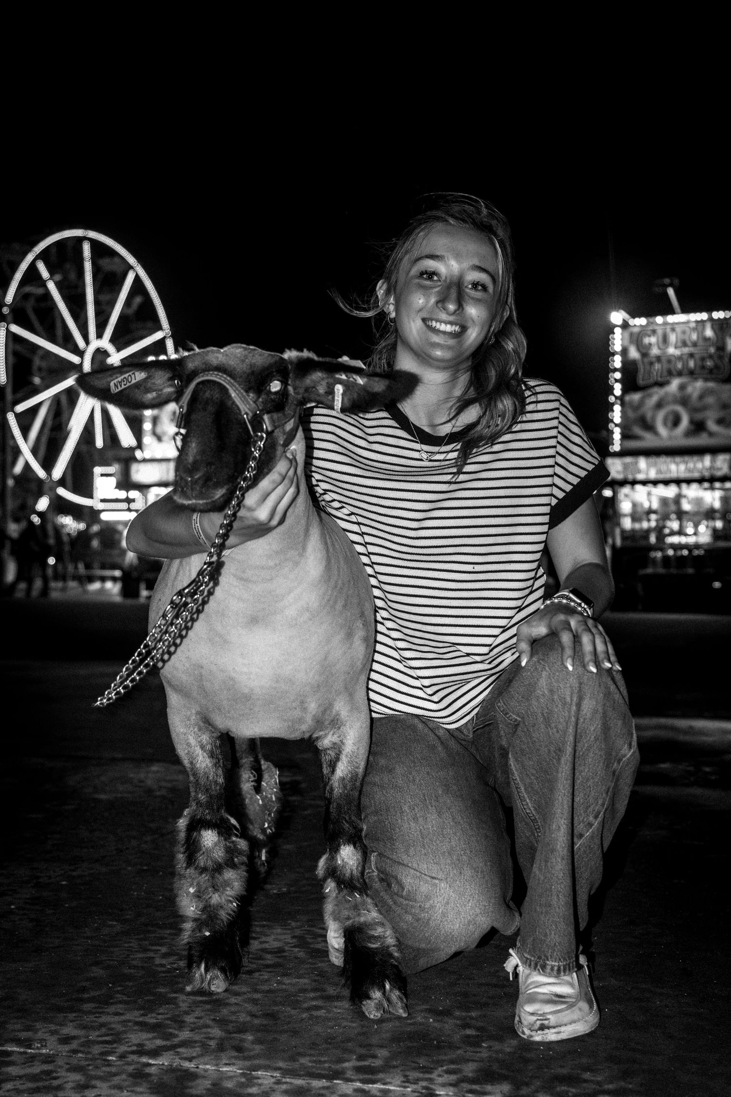 Black and white photo of a girl kneeling with a goat at a county fair with Ferris wheel background.