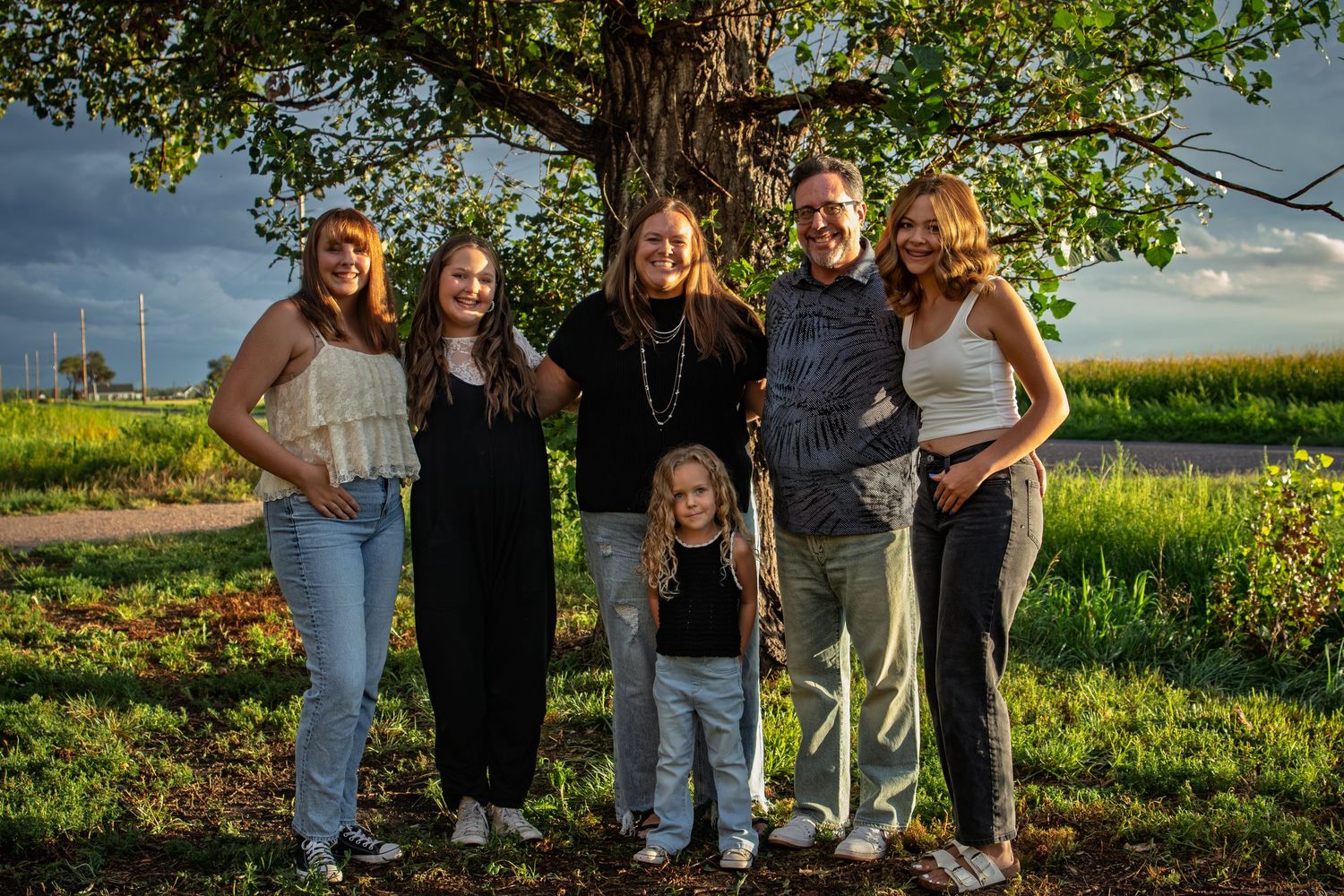 Family of six poses together under a large tree in a lush green field at golden hour.