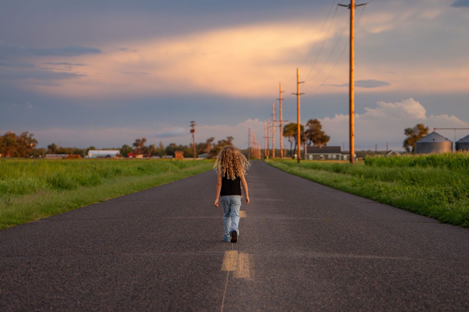 Young girl with curly hair walking away down a rural road at colorful sunset.