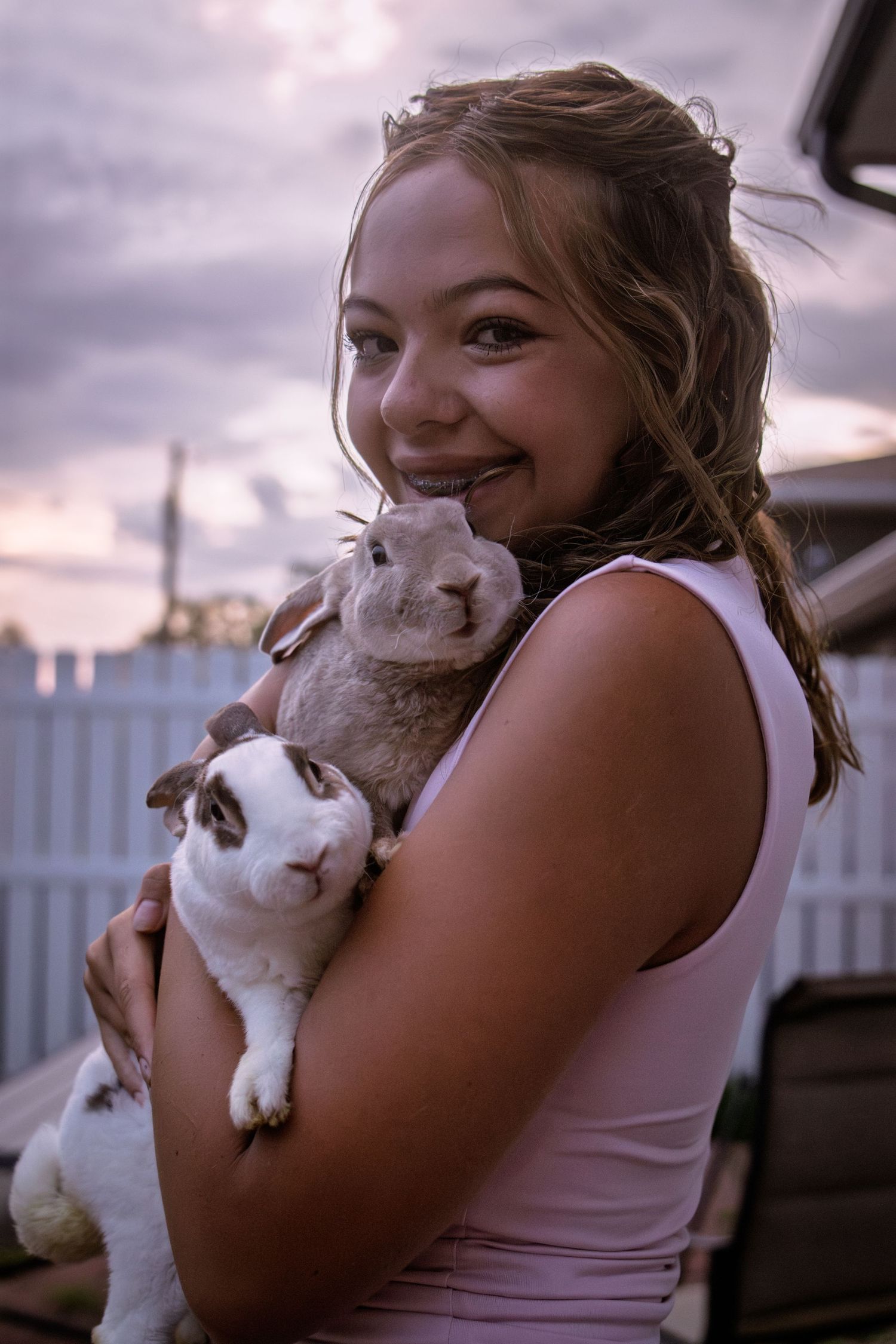 Happy young girl hugging two rabbits close to her chest under a dramatic cloudy sunset sky.