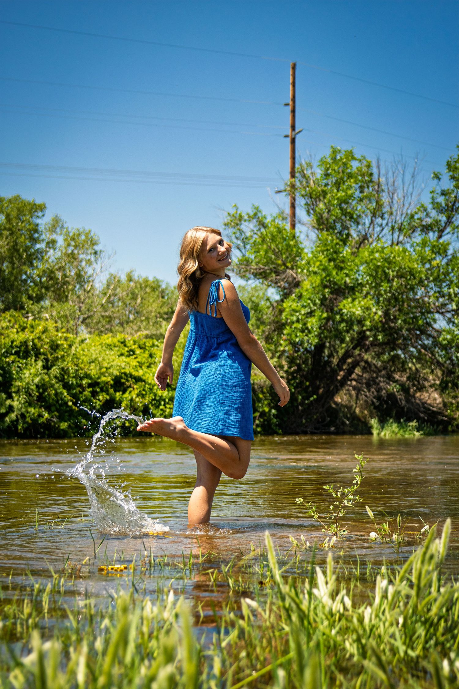 Blonde woman in blue dress splashing water in a river surrounded by green trees.