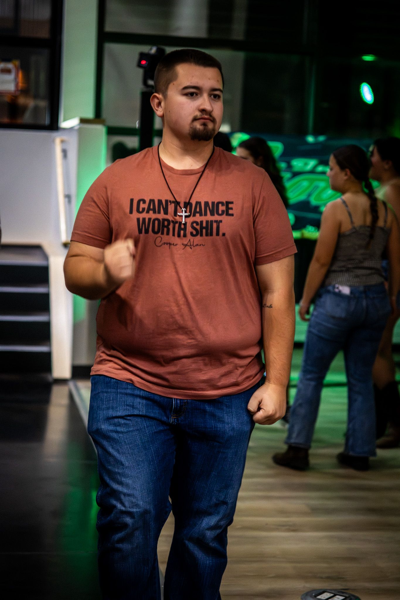Man wearing I Can't Dance Worth Shit t-shirt dancing at a lively indoor event with green lighting.