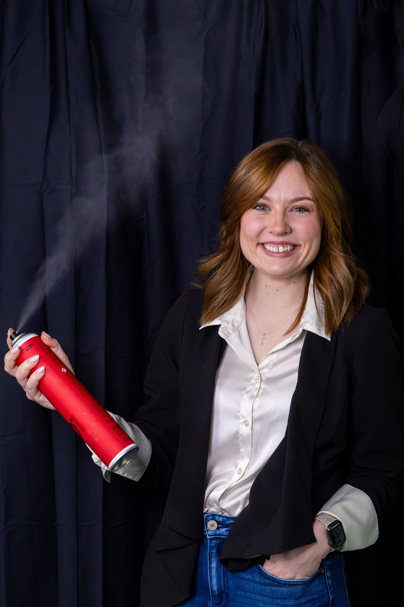 Smiling redhead woman in blazer using red hairspray can against dark curtain backdrop.