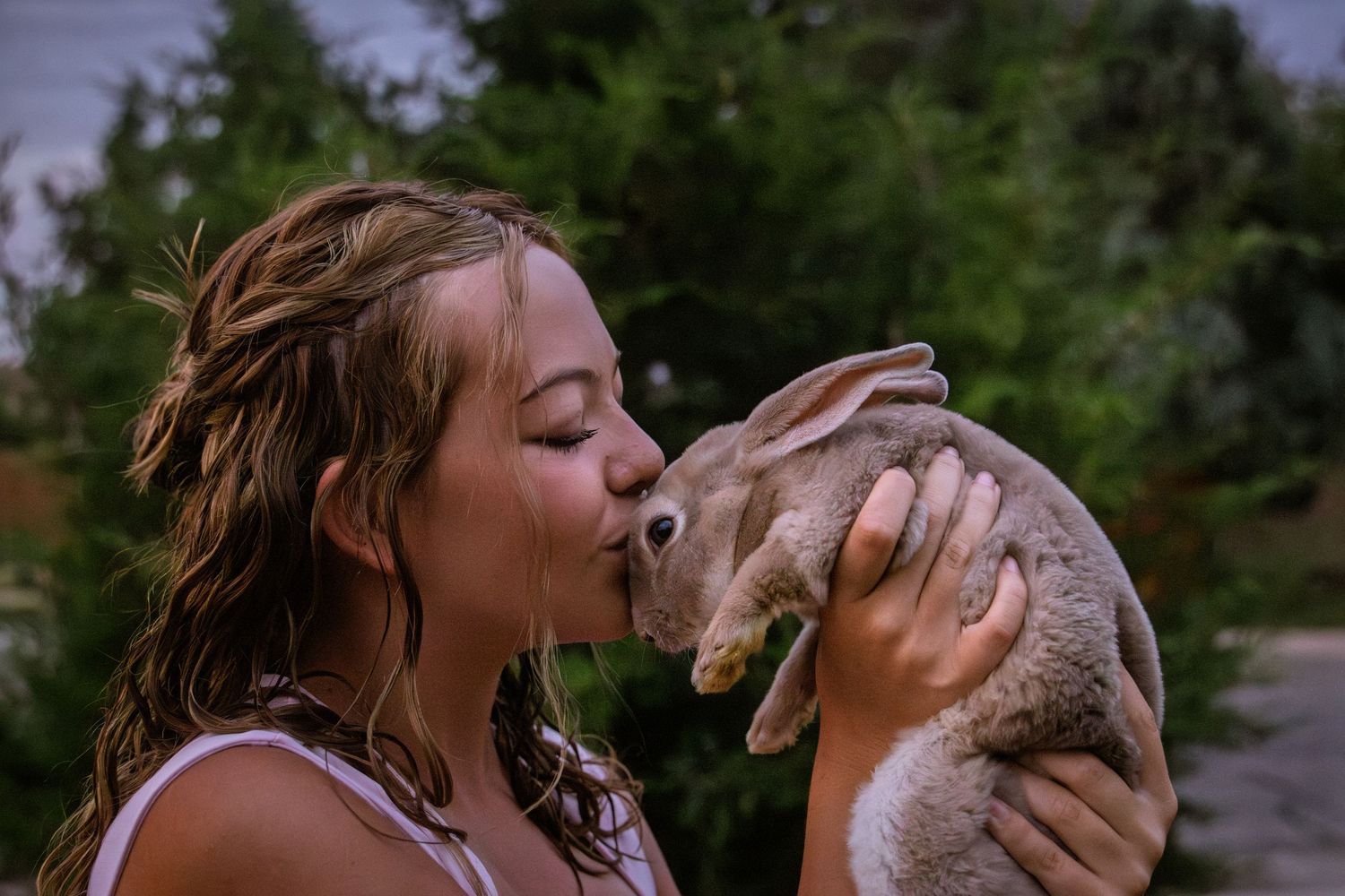 Young girl kissing a rabbit outdoors surrounded by lush green trees in soft evening light.