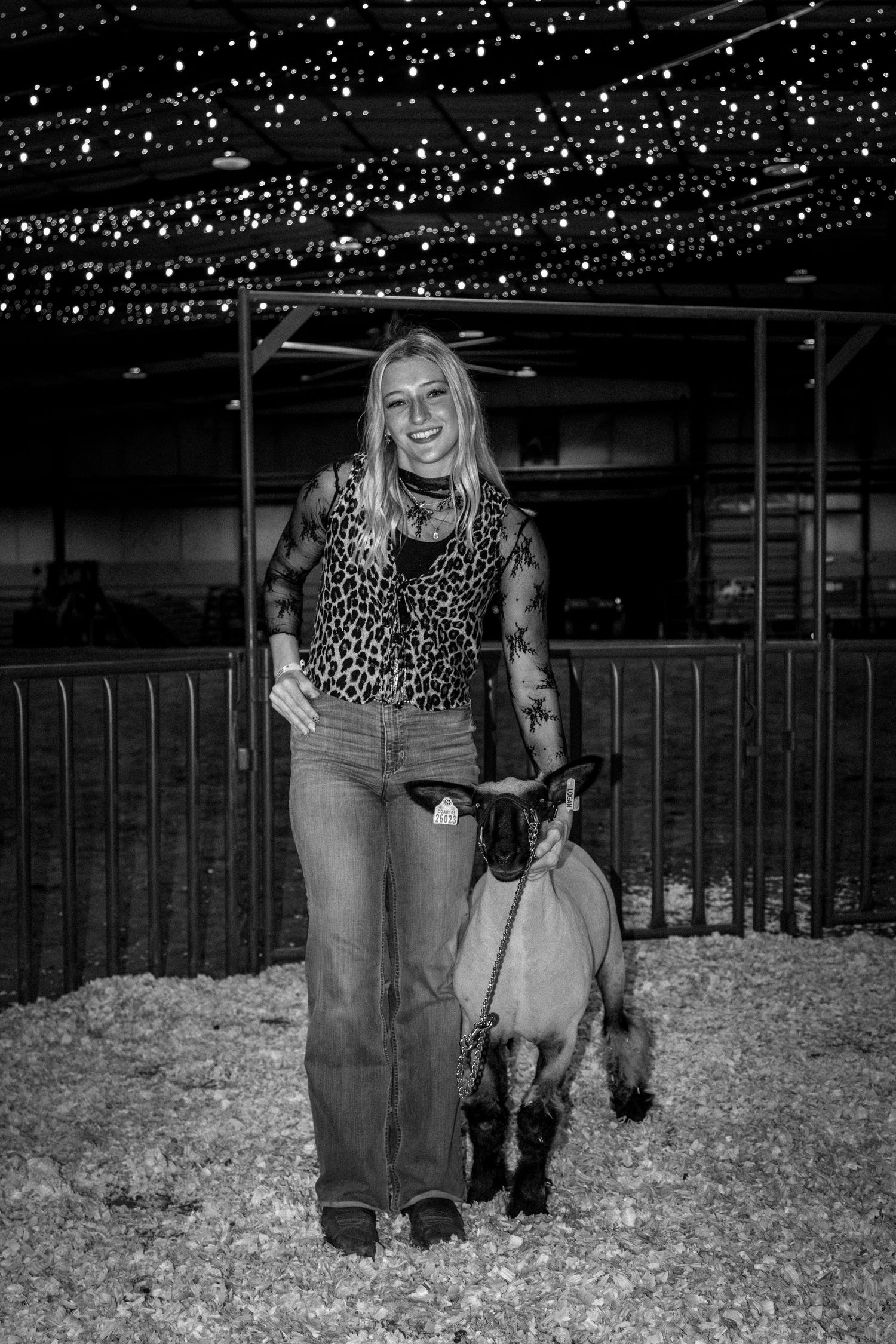 Young woman posing with a goat on a leash inside a livestock barn decorated with string lights.