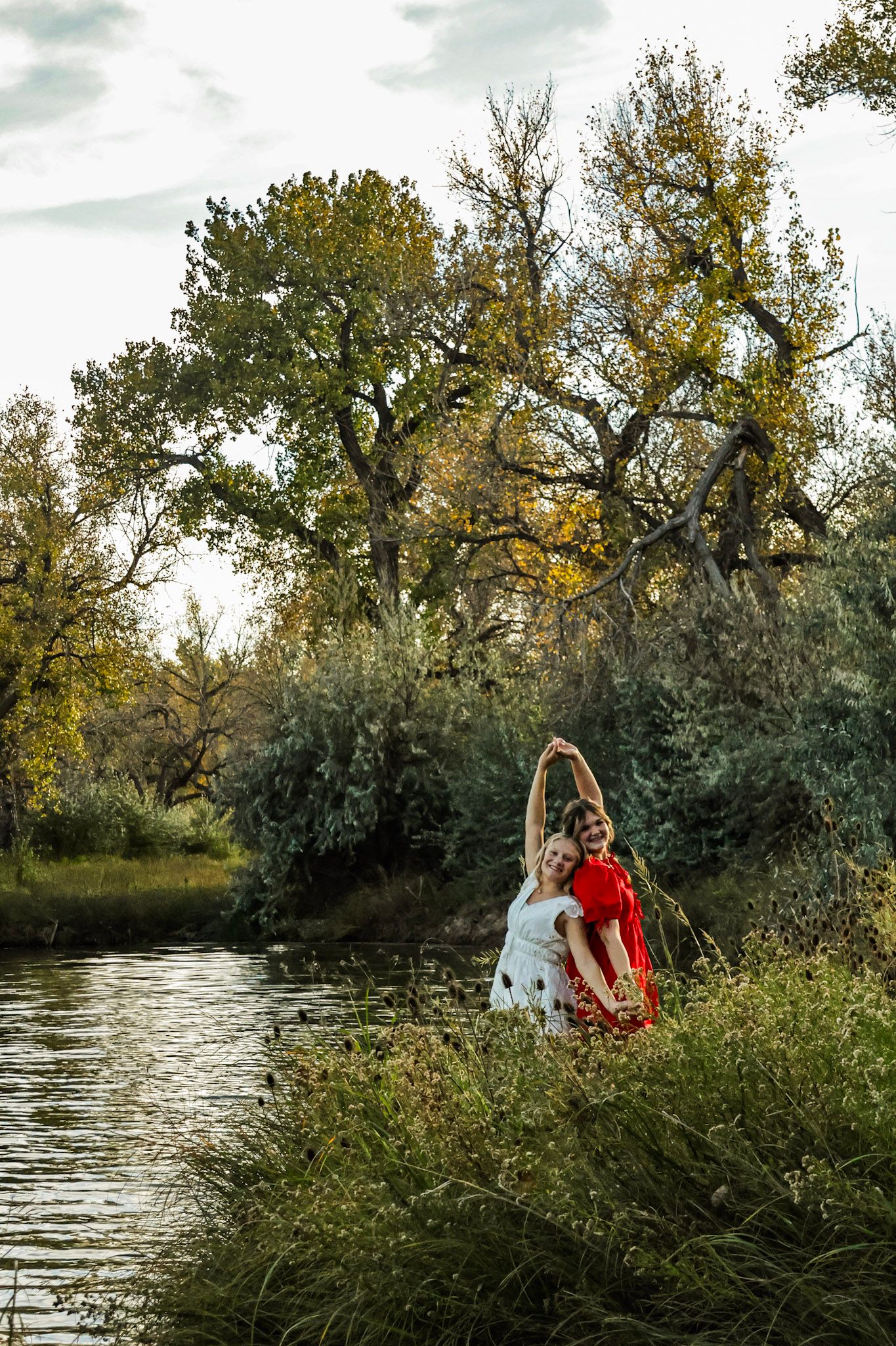 Two girls in white and red dresses dance joyfully together beside a peaceful autumn riverside.