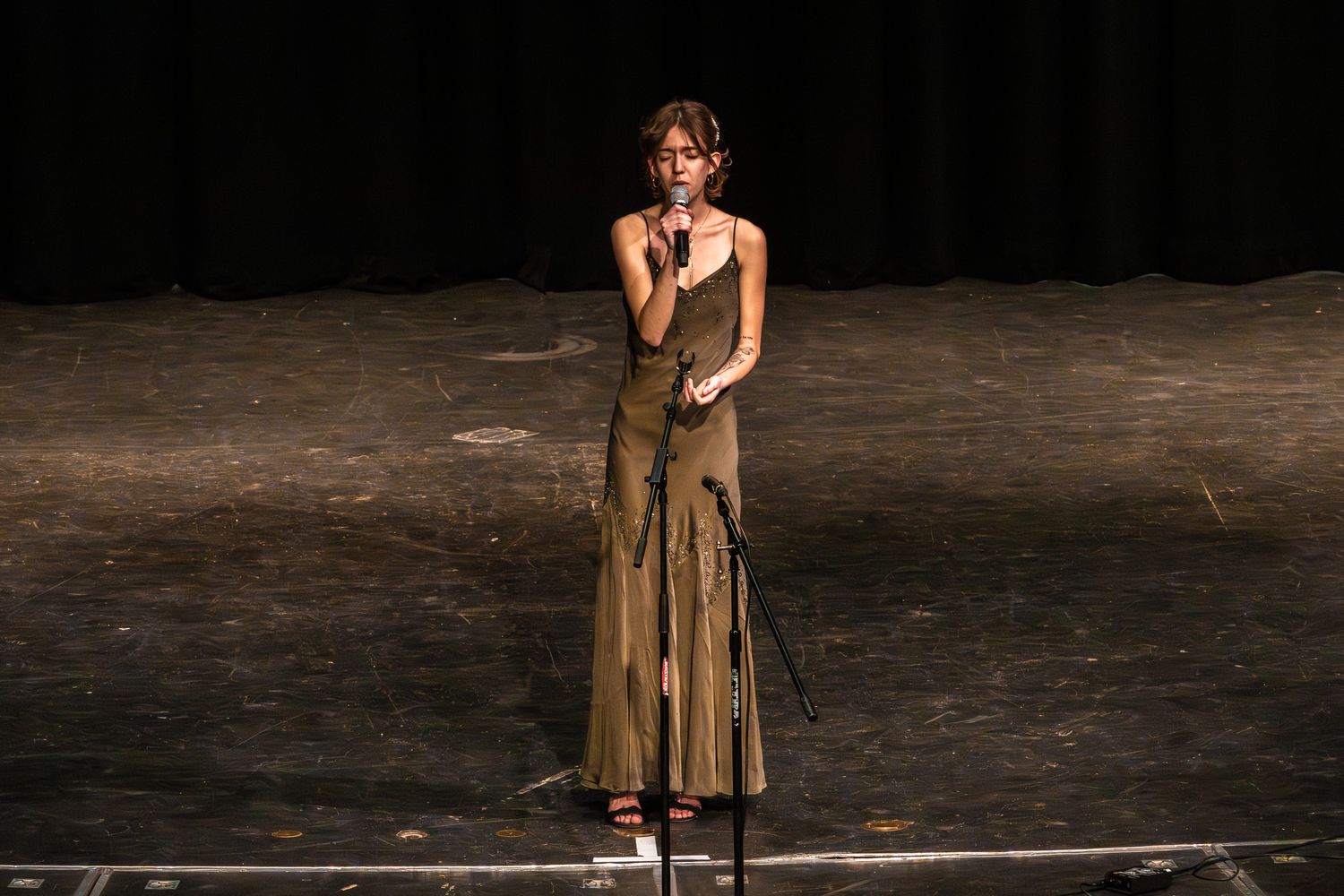Female singer performing on stage in elegant gold gown holding microphone at live event.