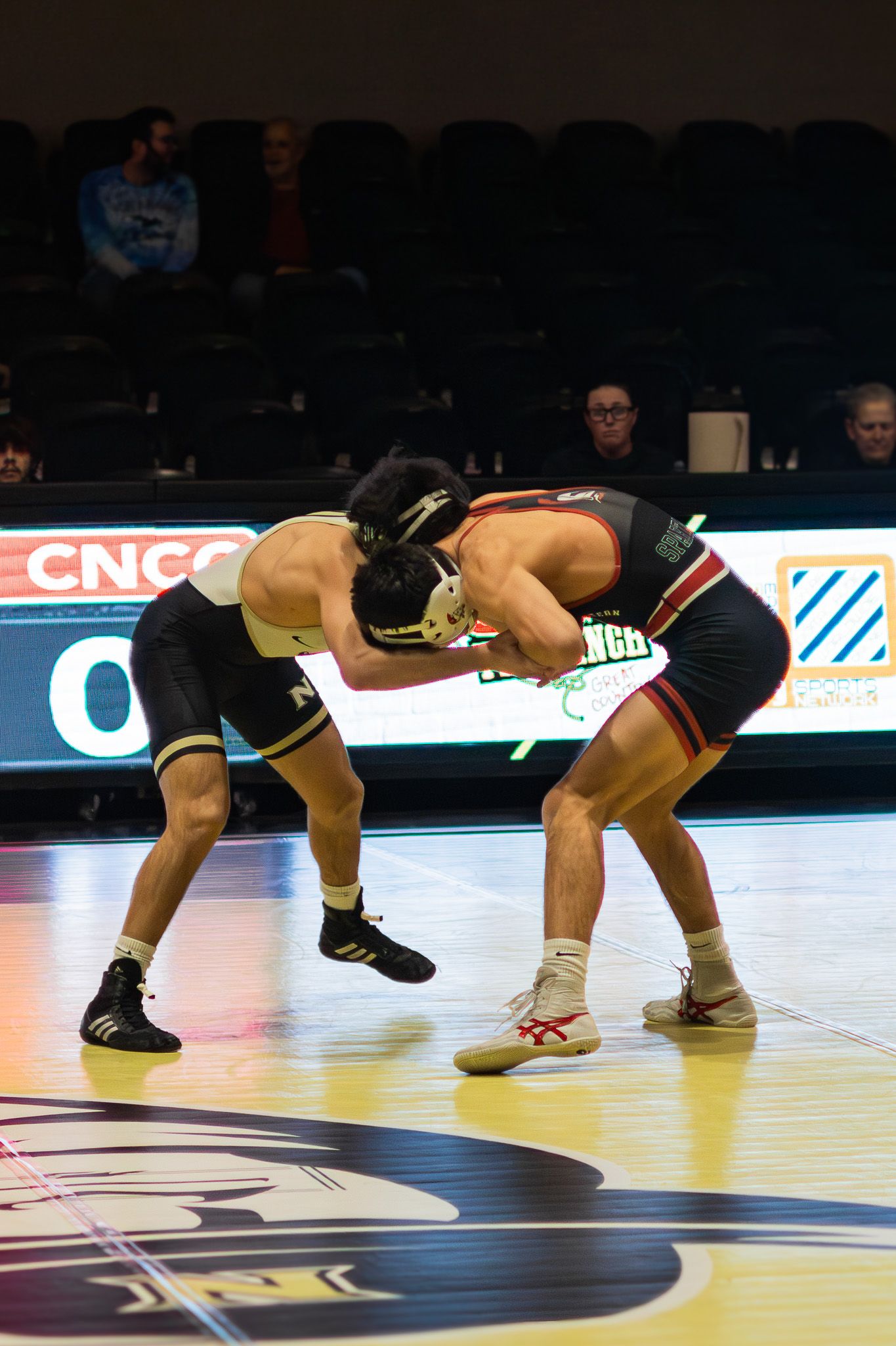 Two college wrestlers in black and red singlets engage in a standing grapple during a match.