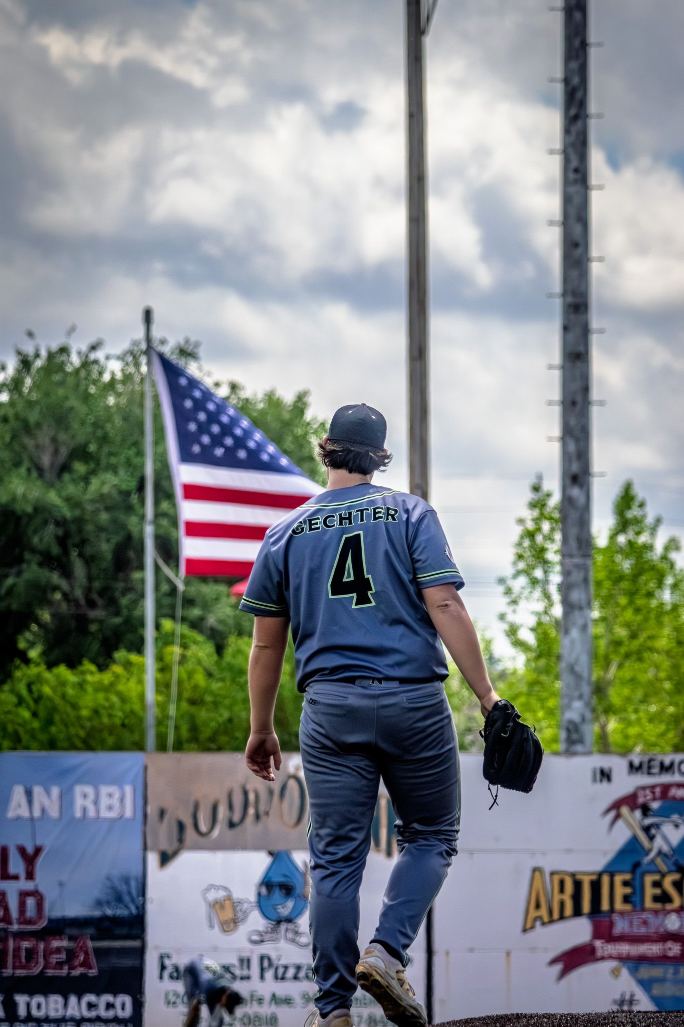 Baseball player Lechtery number 4 walks toward an American flag flying at a baseball stadium.