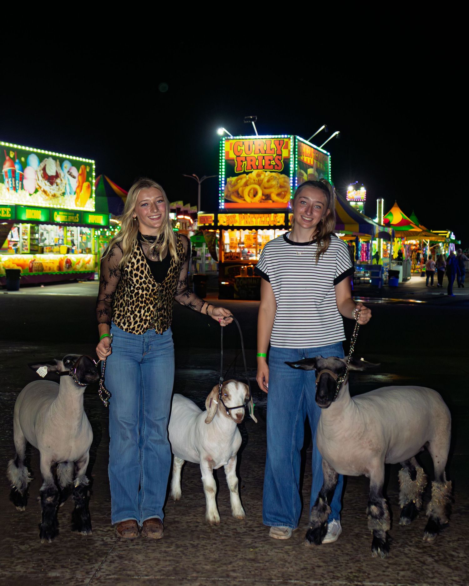 Two girls with sheep and a goat at a county fair at night with colorful carnival lights in background.