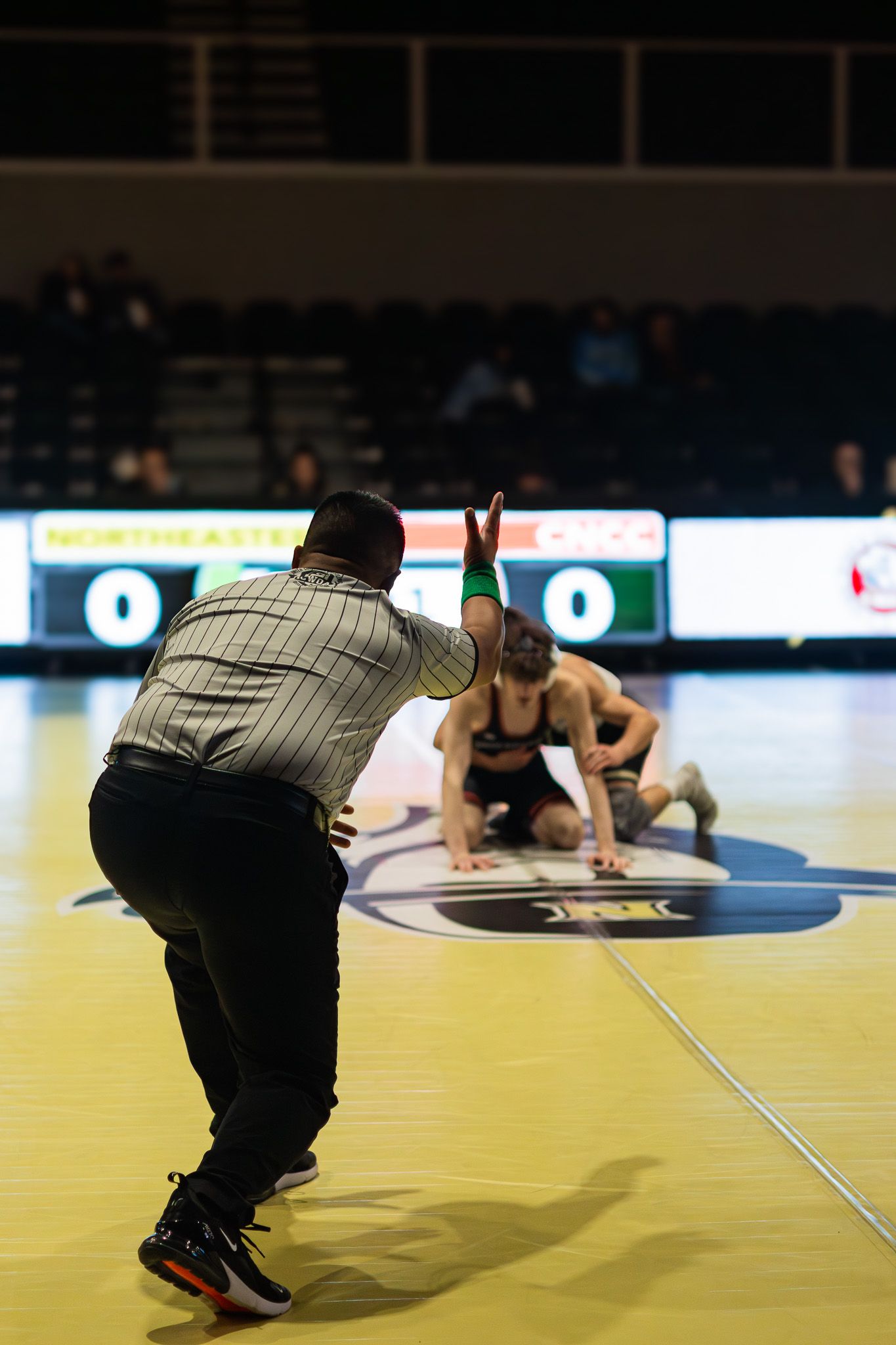 Wrestling referee signals points as two competitors grapple on the mat during an indoor tournament.