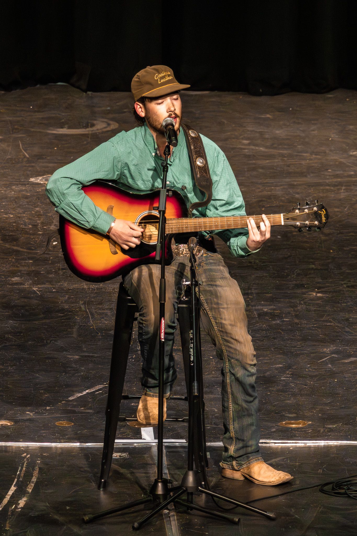 Male musician playing acoustic guitar on stage wearing green shirt and brown cap at live performance.