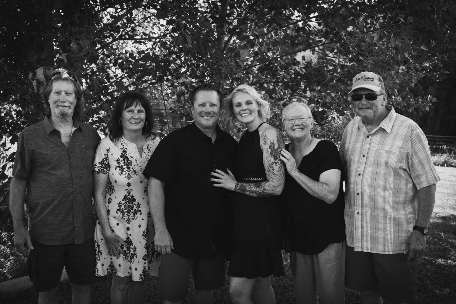 Black and white photo of six family members smiling together outdoors beneath leafy trees.