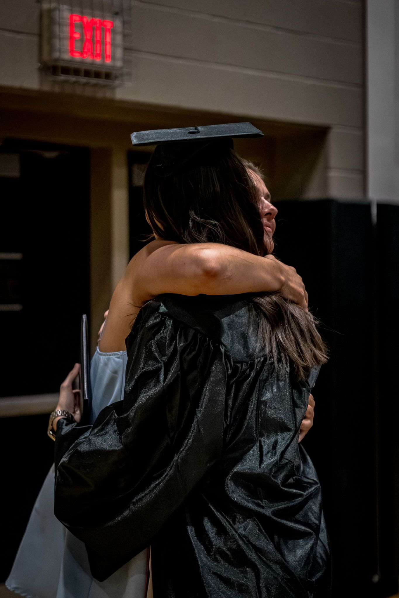 Two people sharing an emotional hug at graduation ceremony near illuminated exit sign.