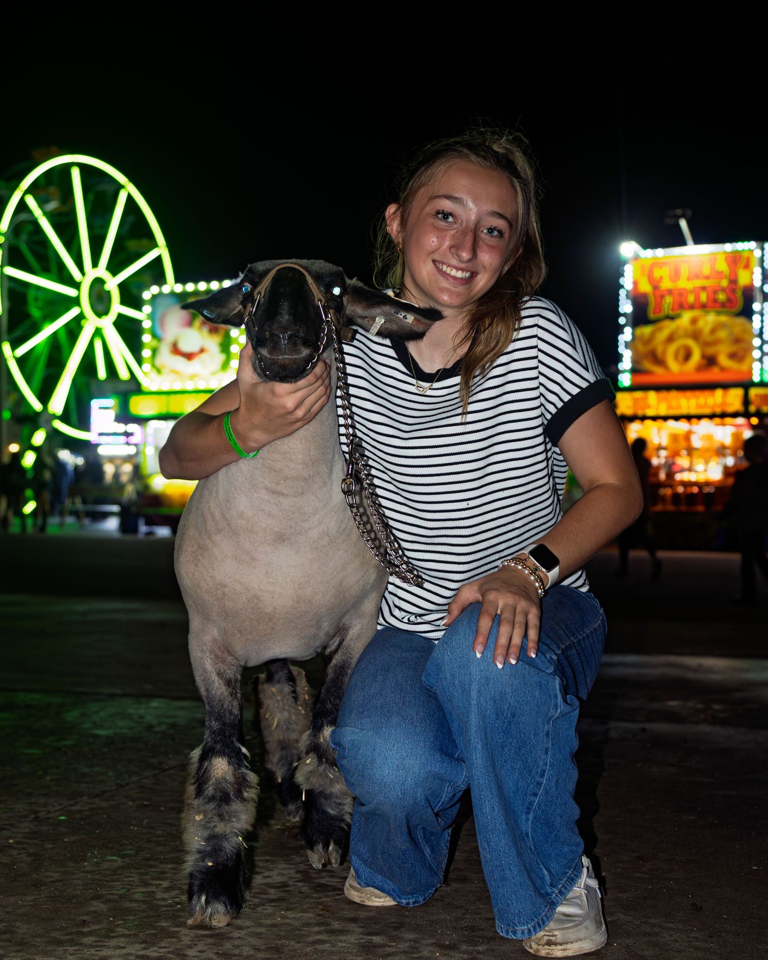 Smiling young woman kneels with grey show lamb at night with lit county fair rides in background.