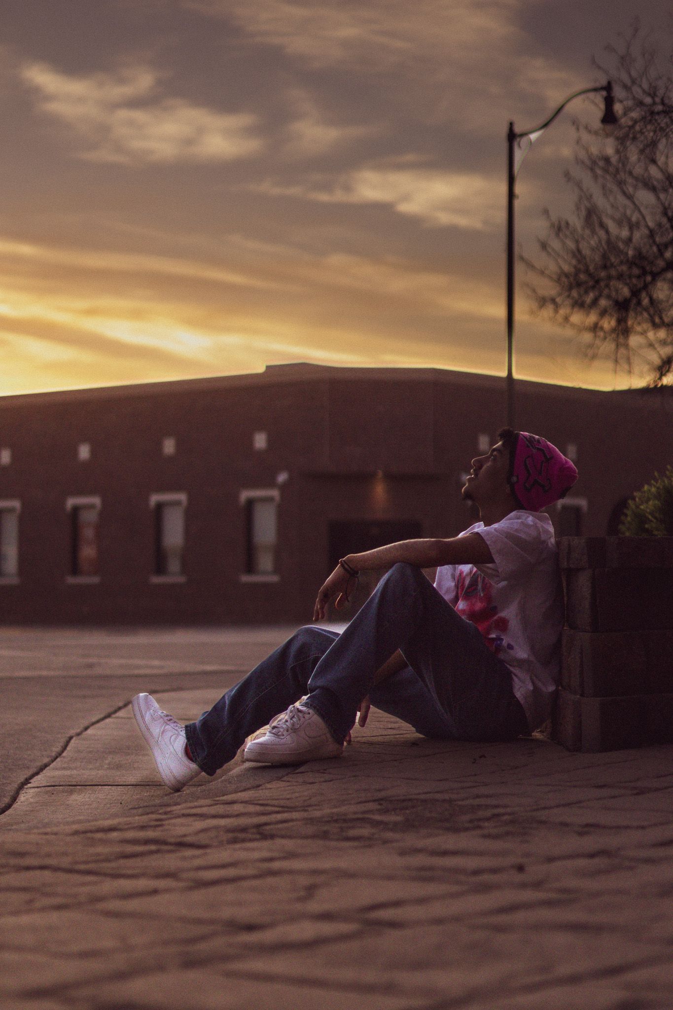 A person in a pink beanie and white tee sits on a brick ledge gazing at a golden sunset sky.