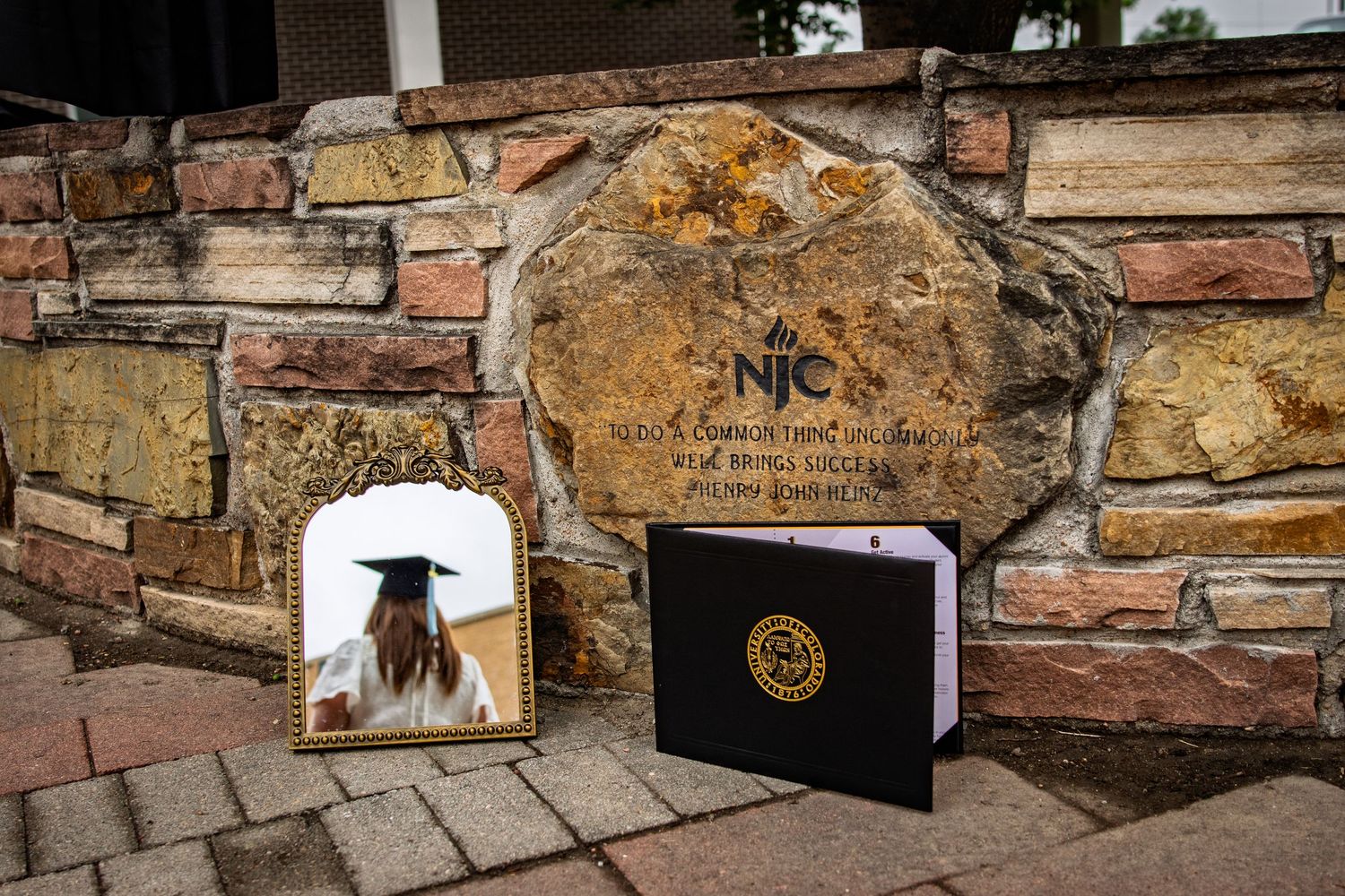 Graduation photo and diploma displayed against NNC college stone wall marker.