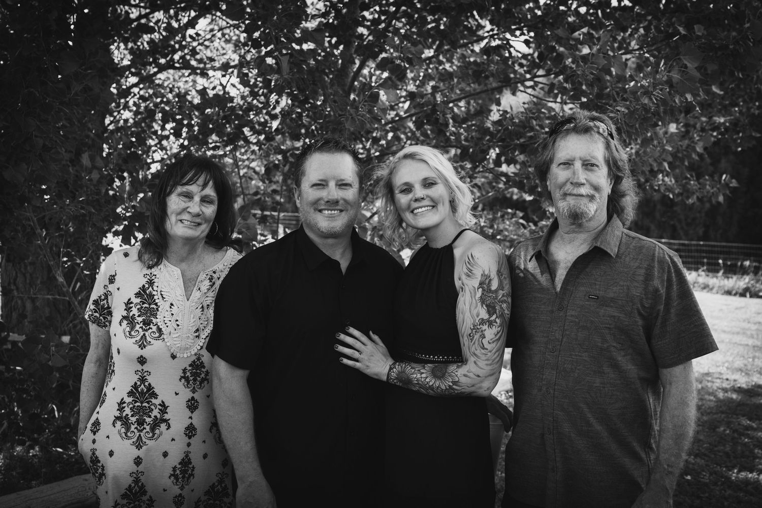 Black and white portrait of four family members smiling together outdoors under leafy trees.