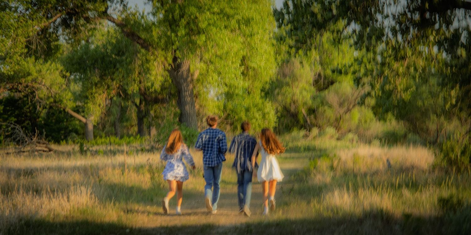 Four people walking together through a lush green meadow with large trees in warm sunlight.