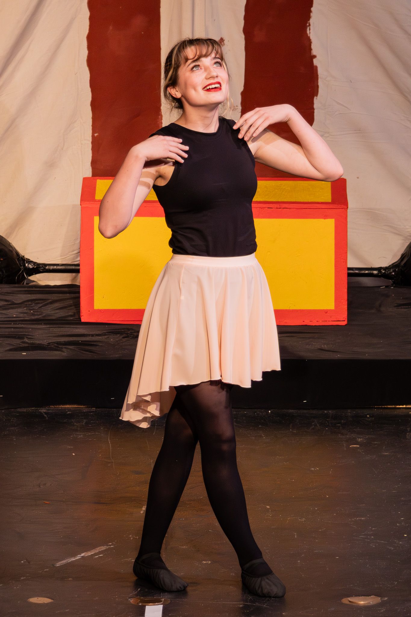 Young woman in black top and beige skirt dances on stage with a colorful circus backdrop.