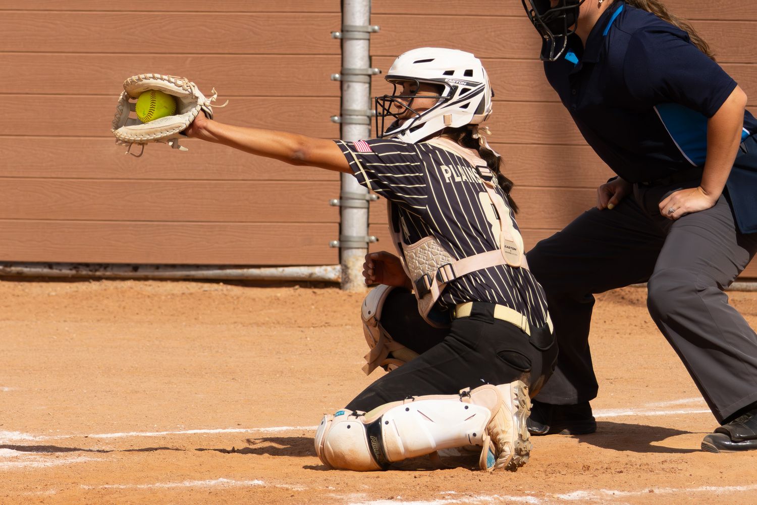 Softball catcher in pinstripe uniform catching ball at home plate with umpire behind.