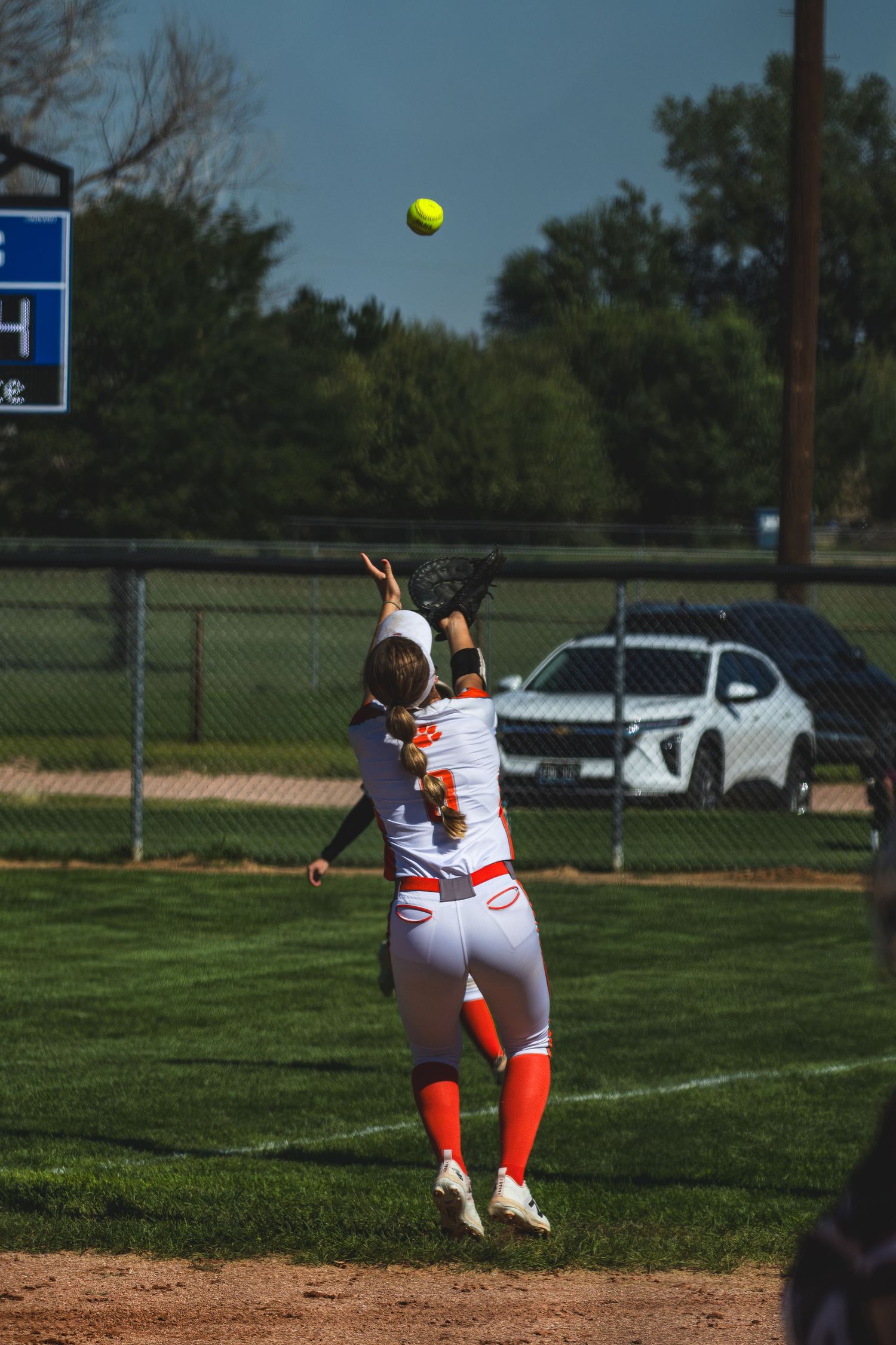 Softball player leaps to catch a fly ball in the outfield wearing orange and white uniform.