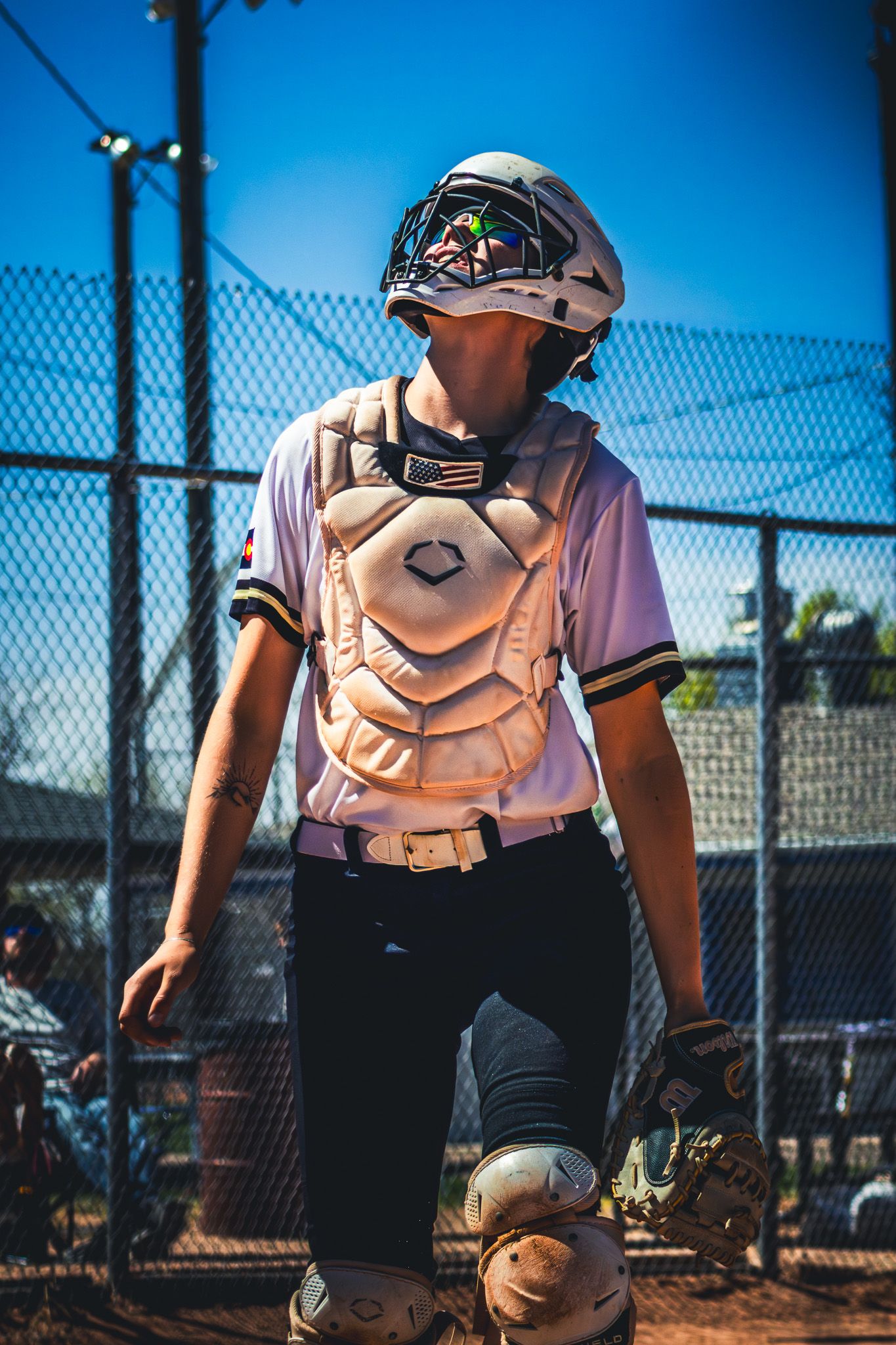 Female softball catcher in full protective gear standing on dirt field under blue sky.