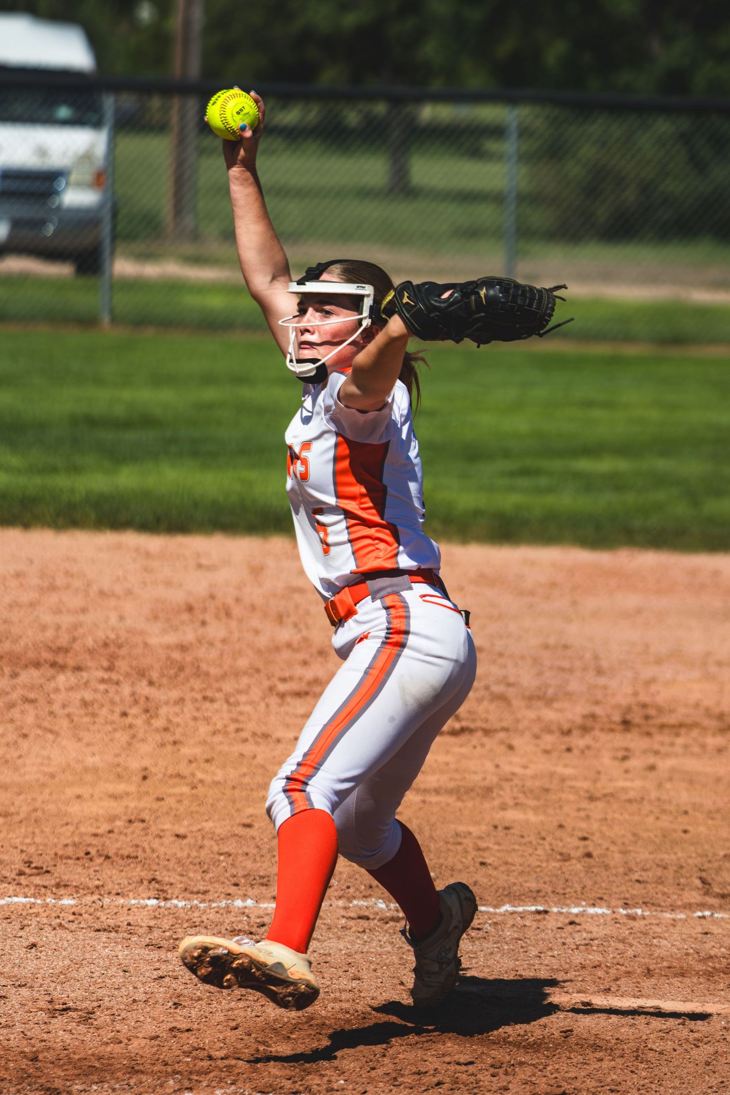 Female softball pitcher winds up to throw a yellow ball during a game wearing orange and white uniform.