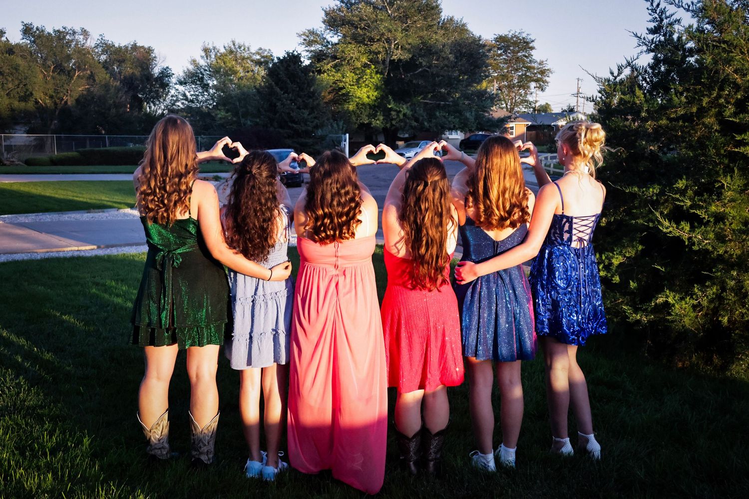 Seven girls in colorful formal dresses stand together making heart shapes with their hands at golden hour.