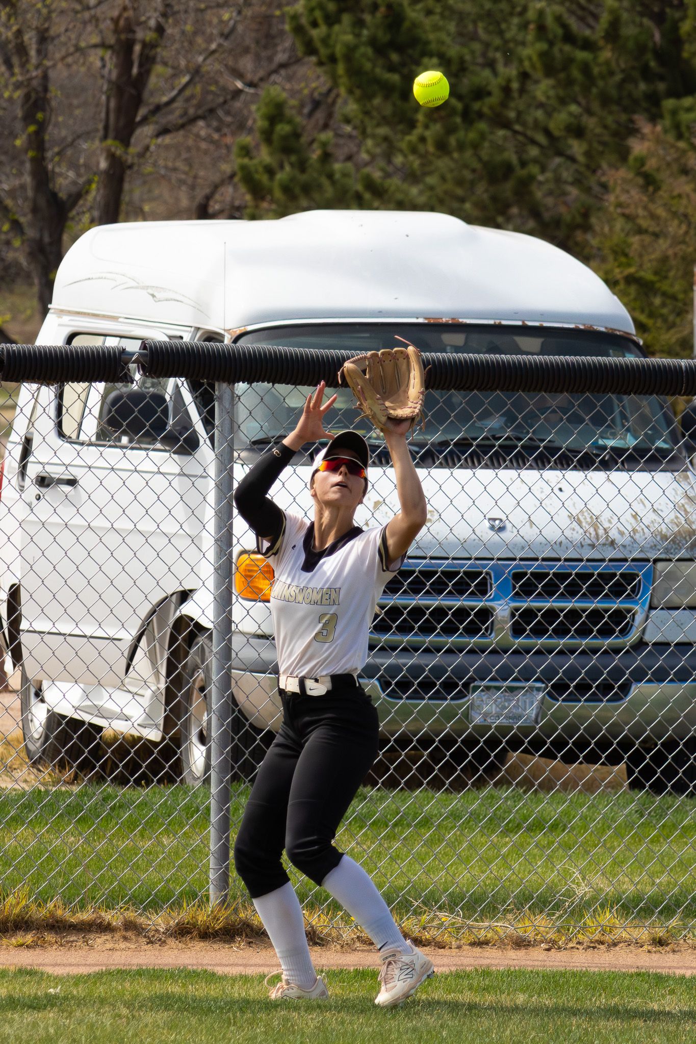 Female softball outfielder reaches up to catch yellow ball near chain-link fence.