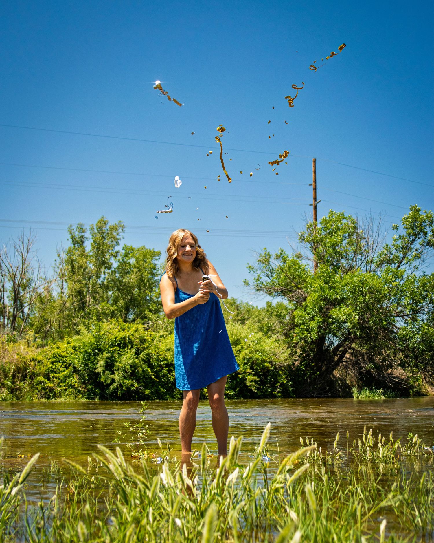 Young woman in blue dress tossing flowers in a shallow river on a sunny day.