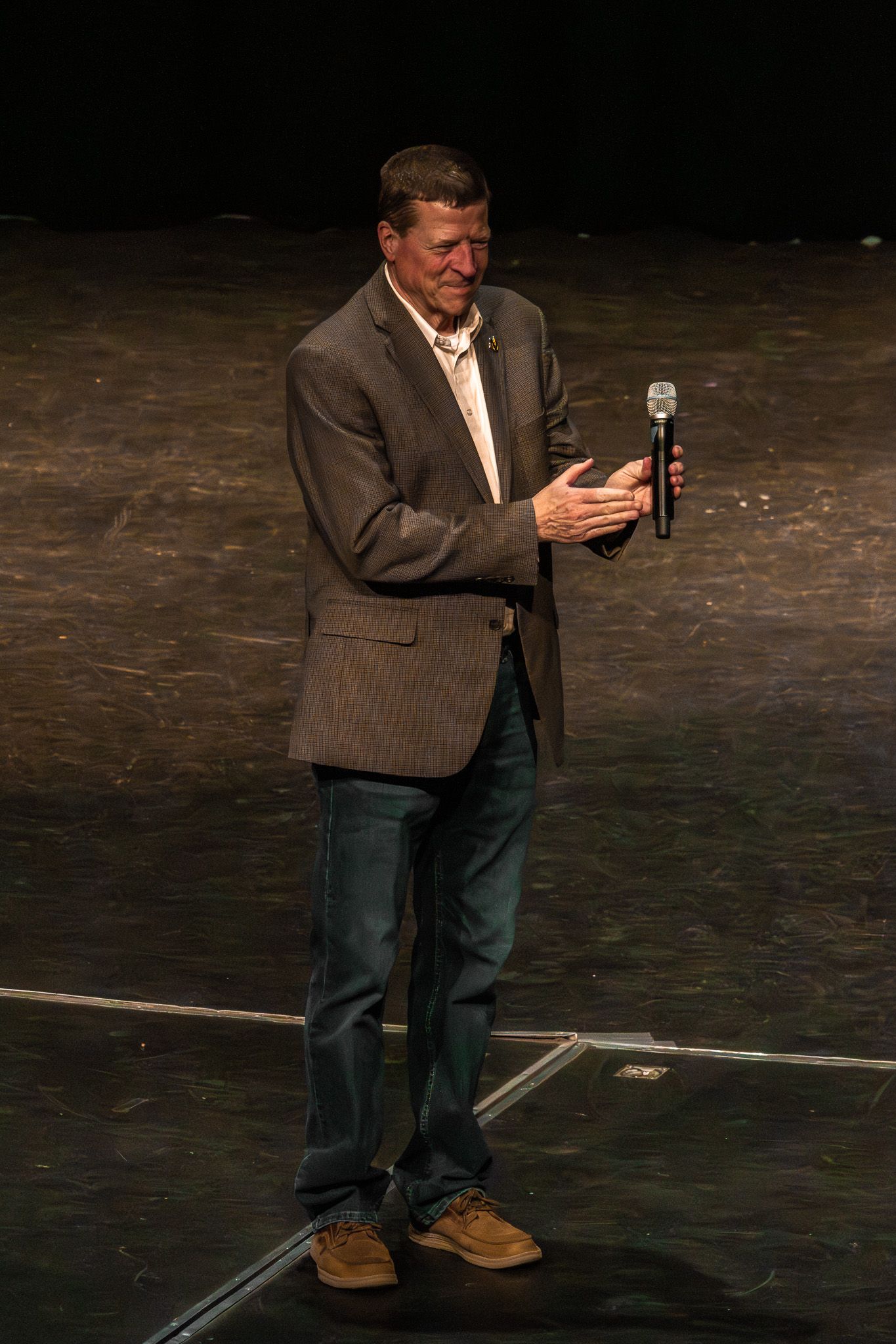 Man in brown blazer and jeans speaks on stage while holding a microphone.