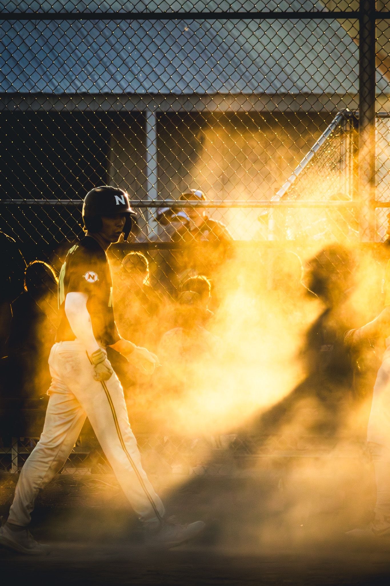 A baseball batter walks through a dramatic cloud of golden dust illuminated by sunset light.