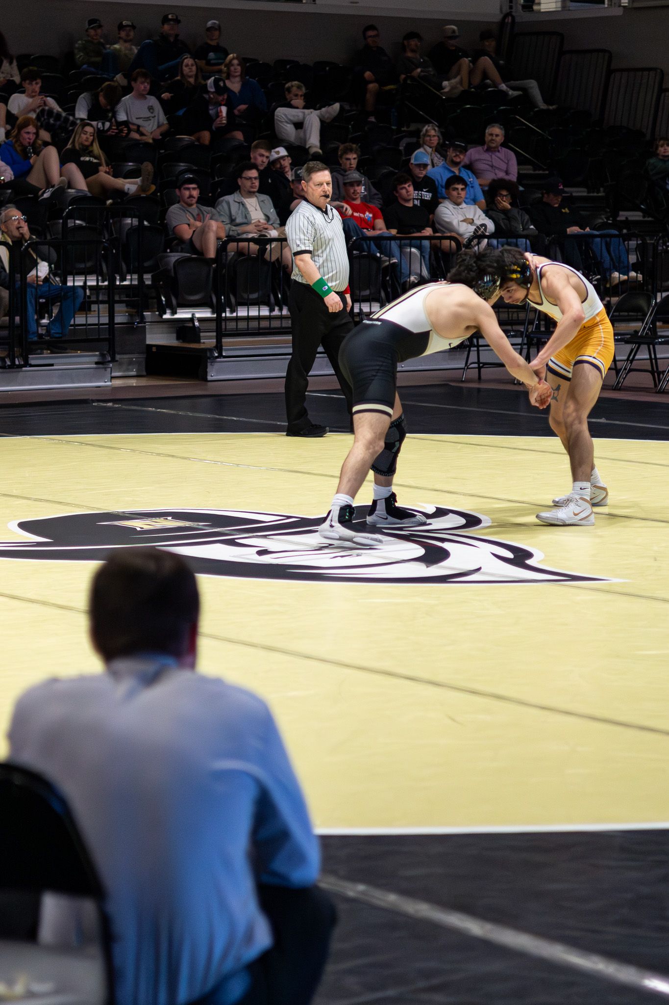 Two college wrestlers in starting position on mat with referee and crowd watching.