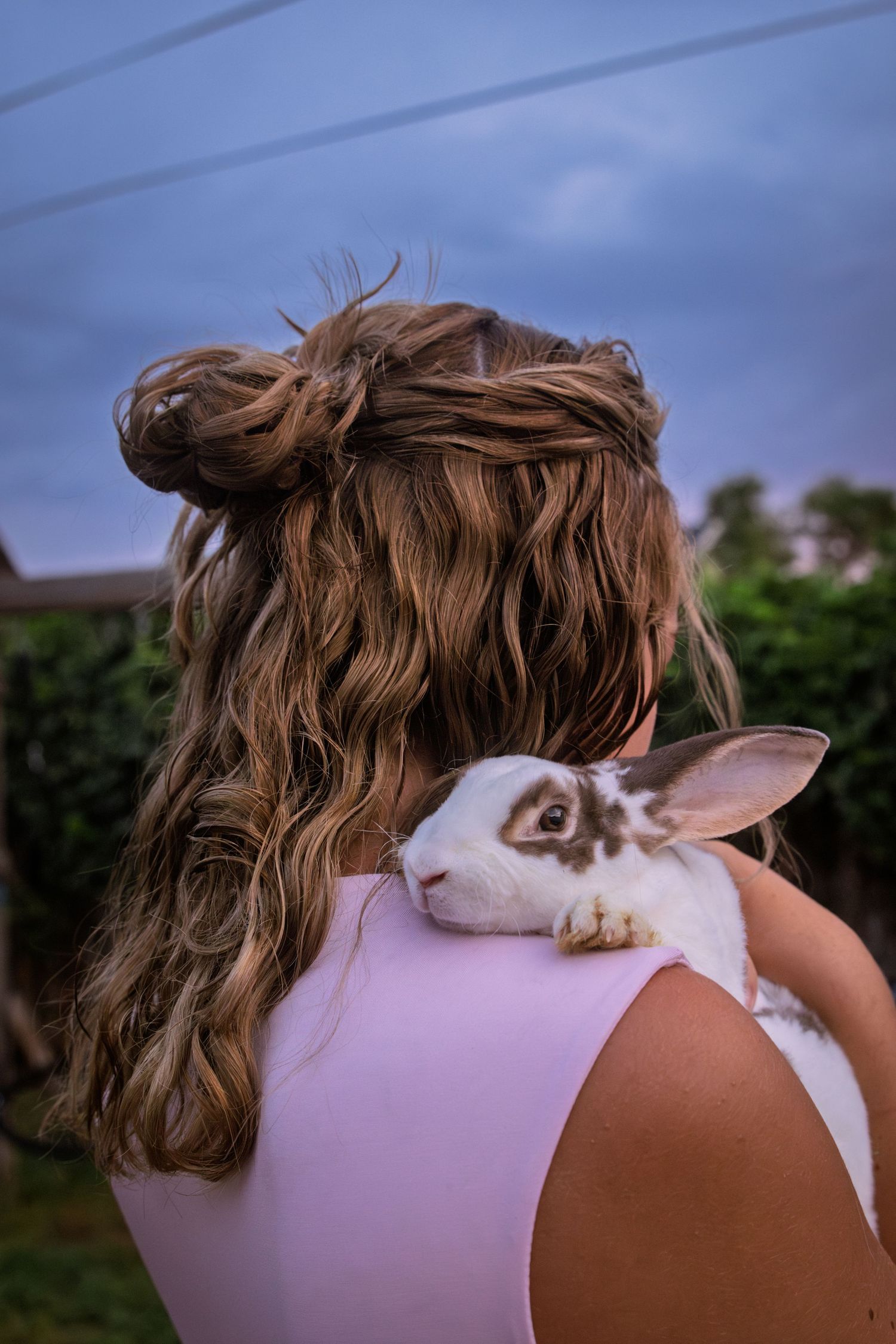 Young girl with curly updo hairstyle holds a white and brown rabbit on her shoulder outdoors.