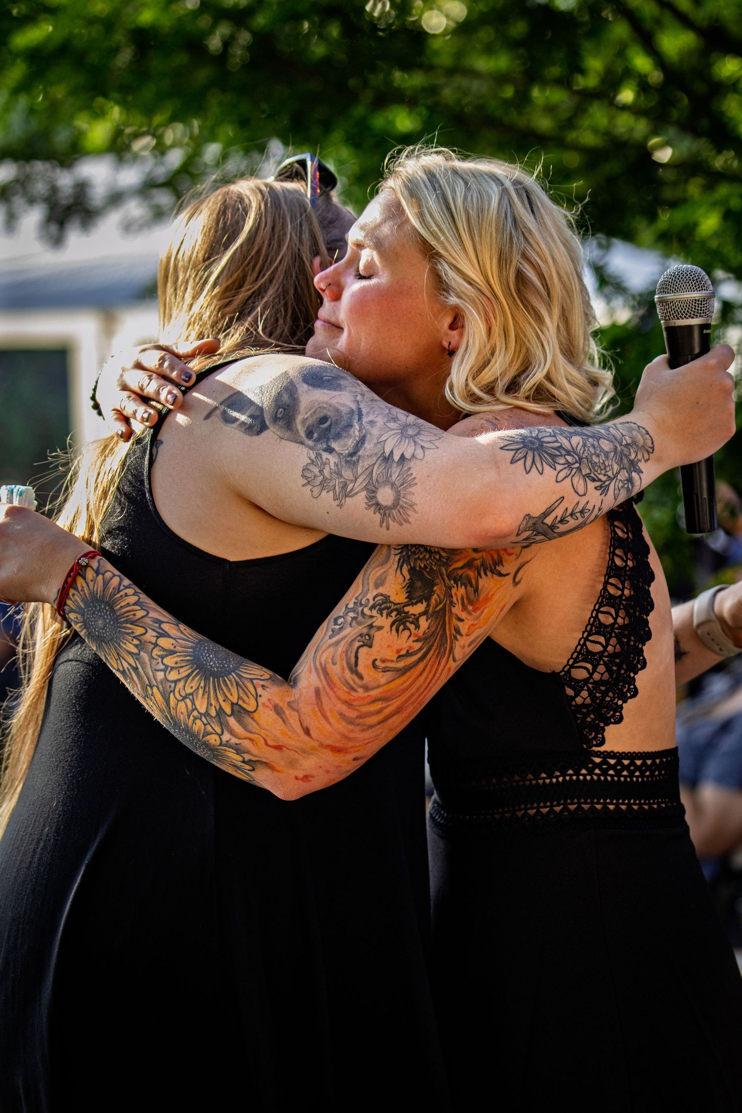 Two tattooed women in black dresses share an emotional hug outdoors at a summer event with microphone in hand.