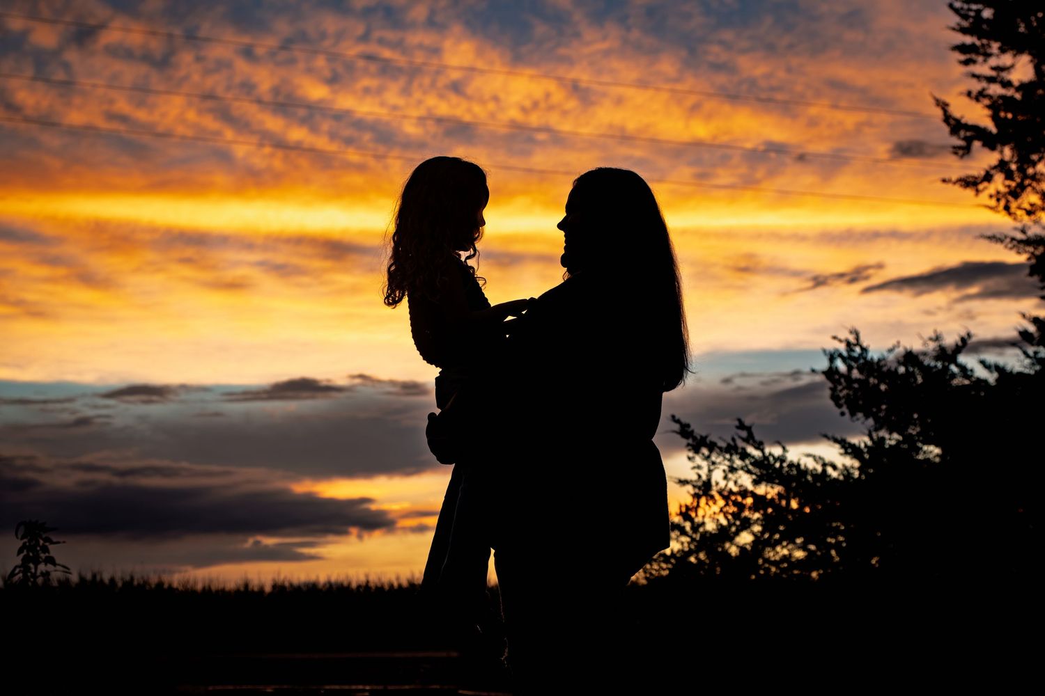 Silhouette of mother holding young child against dramatic orange and yellow sunset sky.
