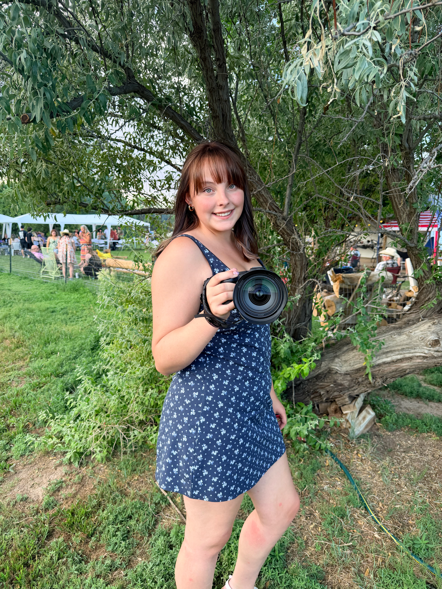 Smiling young woman in floral dress holds a DSLR camera under a willow tree at outdoor event.