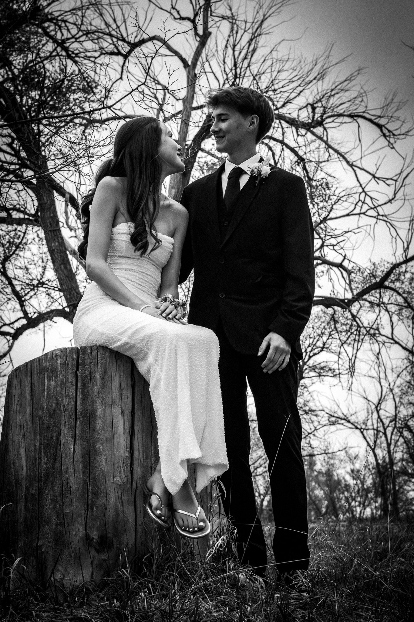 Black and white portrait of a young couple in formal wear near a tree stump in a forest.