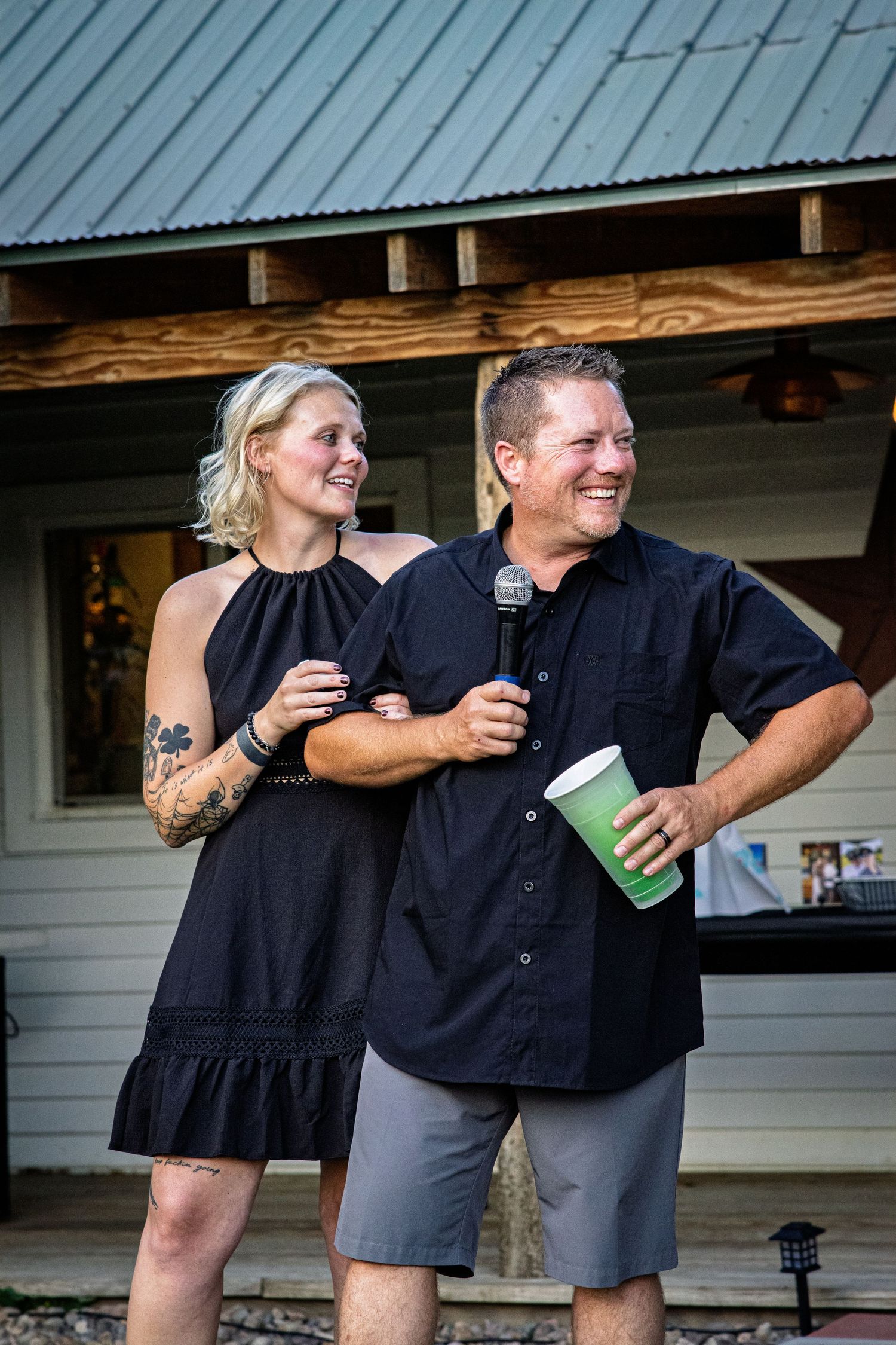 Smiling blonde woman with tattoos and a man holding a microphone and cup speak together at an outdoor event.