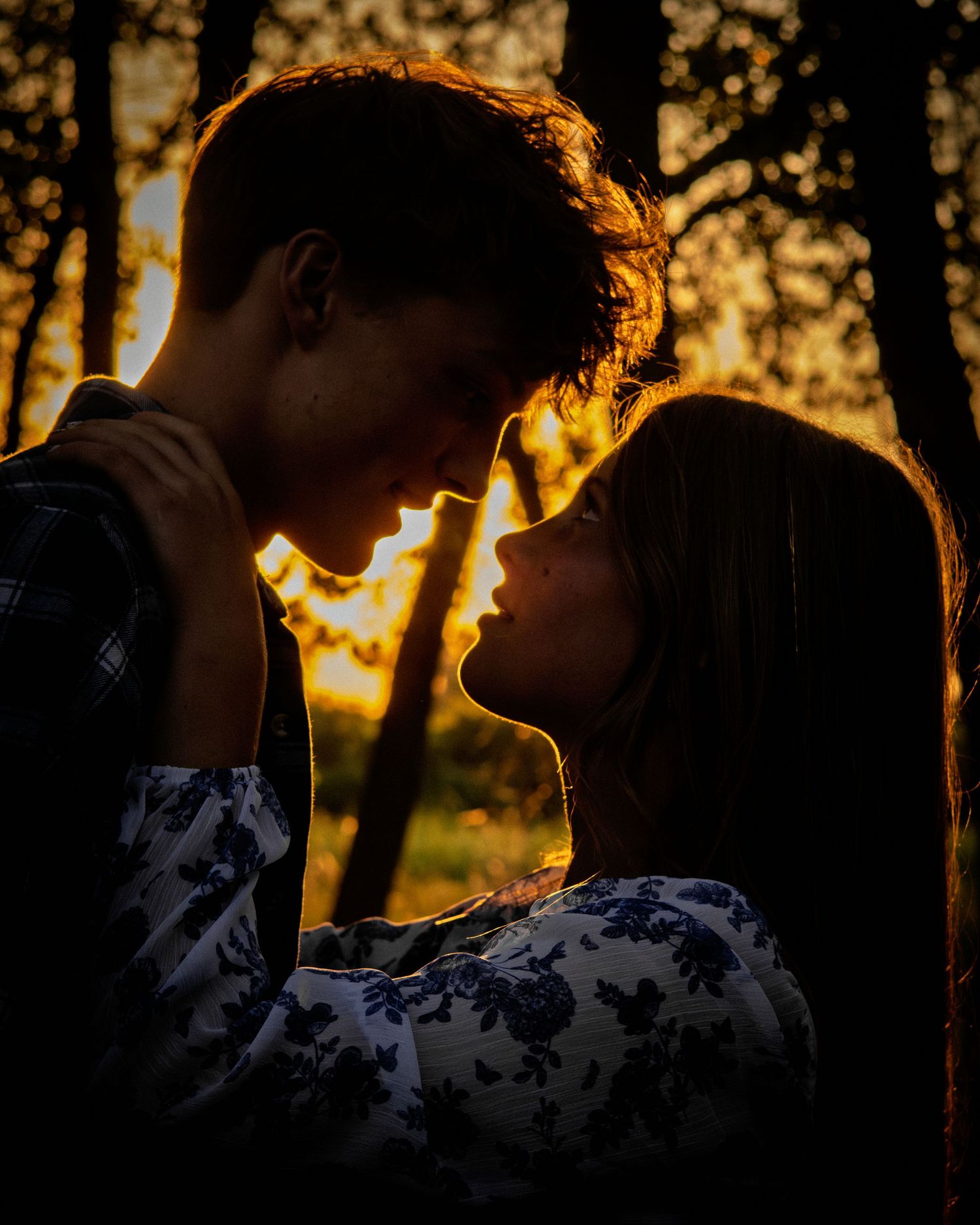 Young couple leaning in close together bathed in warm golden sunset light among trees.