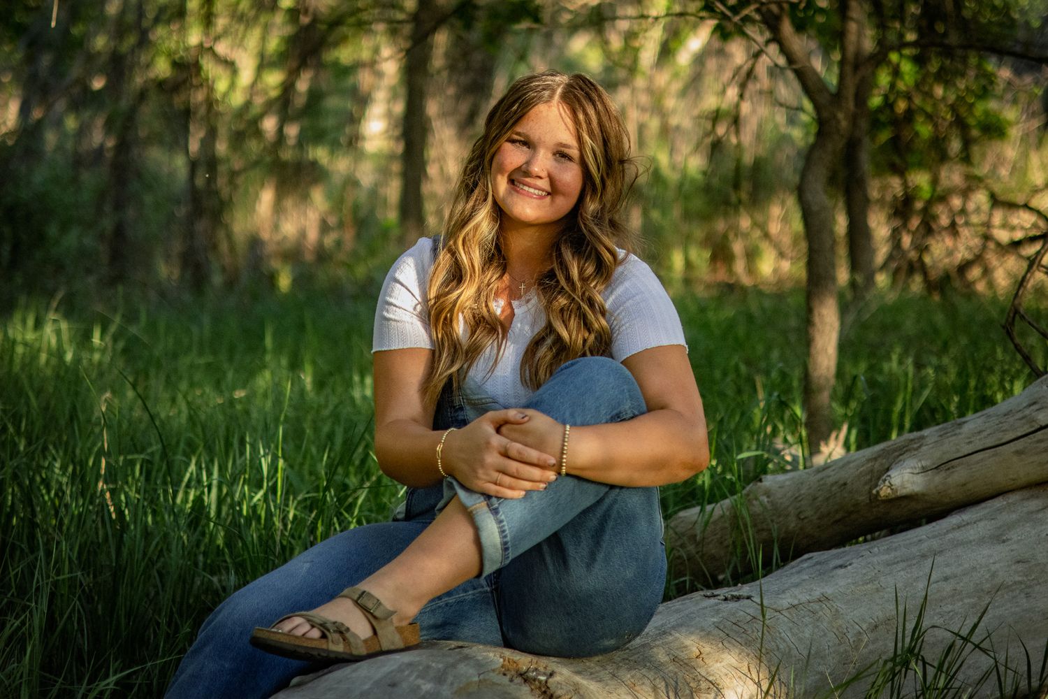 Smiling young woman in white top and jeans sitting on log in lush green forest setting.
