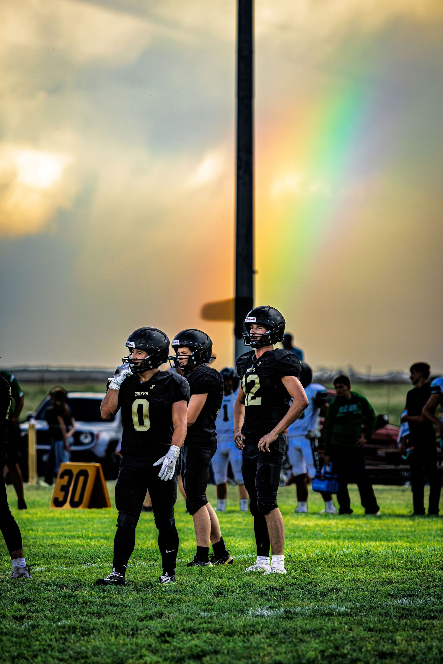 Three football players in black uniforms stand on the field with a vivid rainbow behind them.