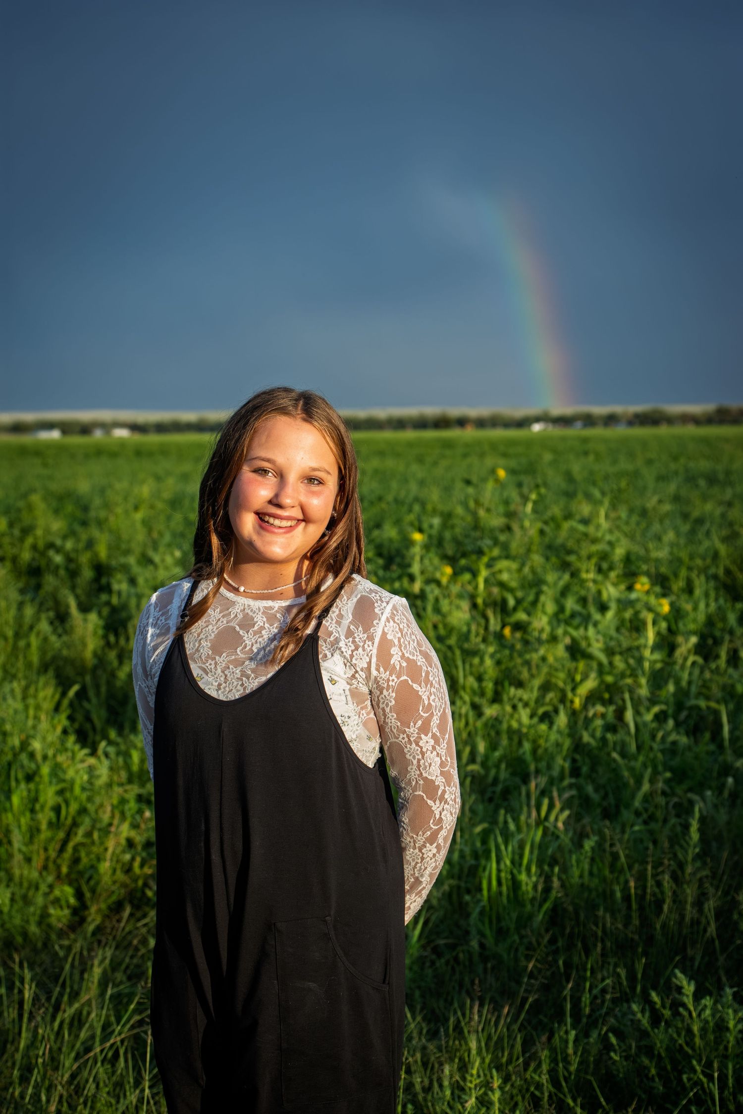Smiling teenage girl in black dress with lace sleeves standing in green field with rainbow in background.