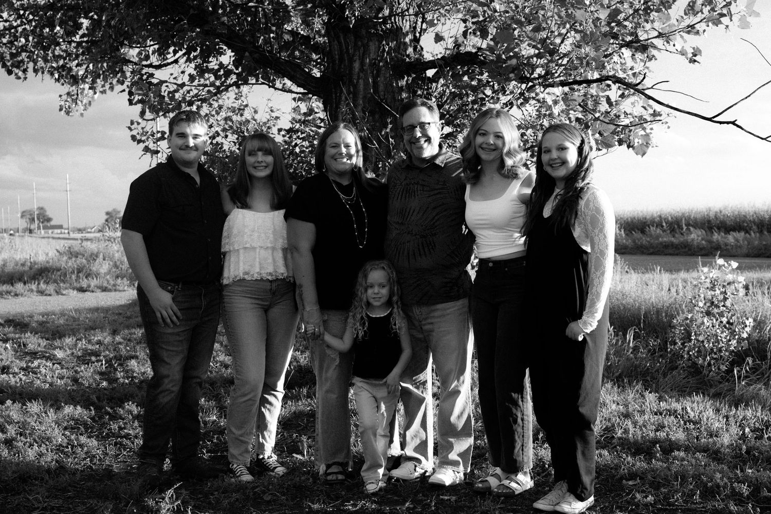 Black and white family portrait of seven people standing together under a large tree outdoors.